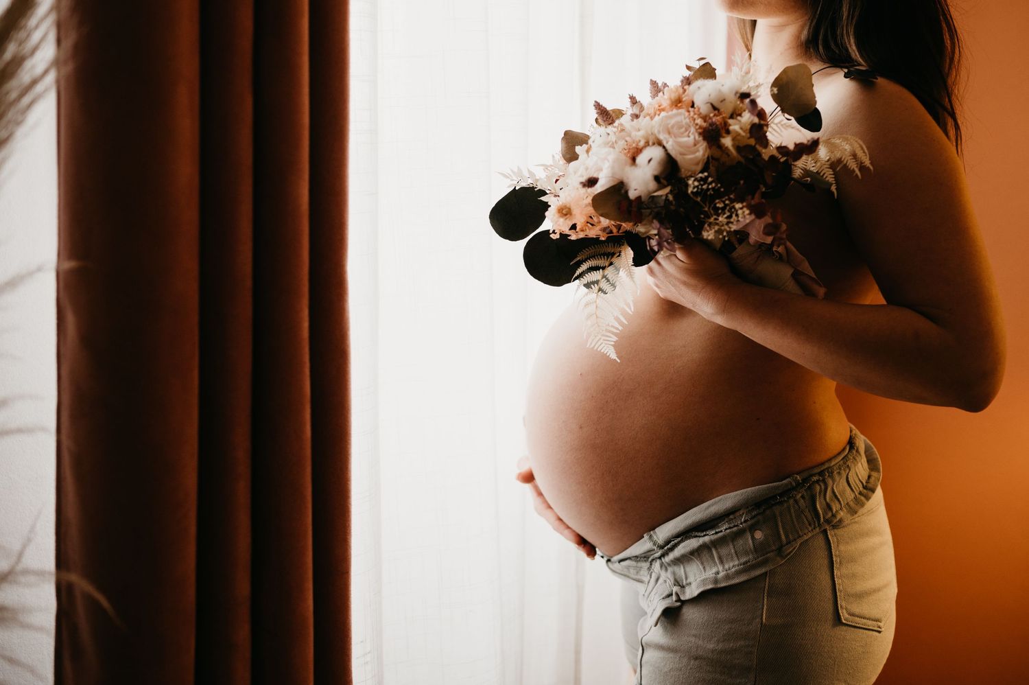 Main de la future maman sur son ventre, un bouquet de fleurs naturelles à la main, séance lifestyle à domicile à Toulouse