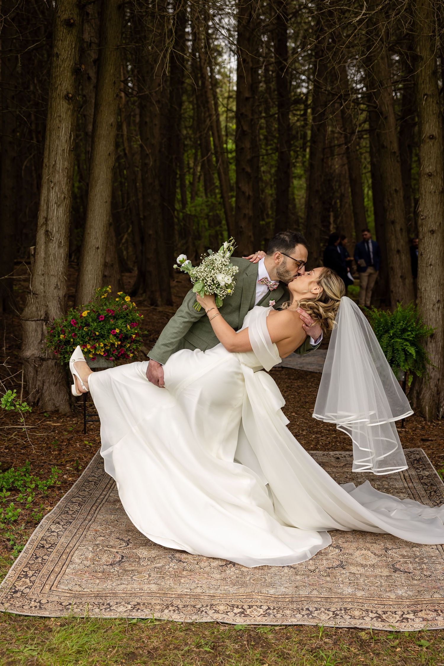 Bride and groom share a romantic dip kiss on a vintage rug surrounded by tall trees in a forest wedding setting.