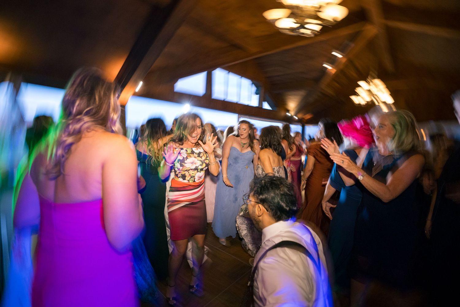 Guests dancing and celebrating at a lively wedding reception with colorful lights in a rustic venue.