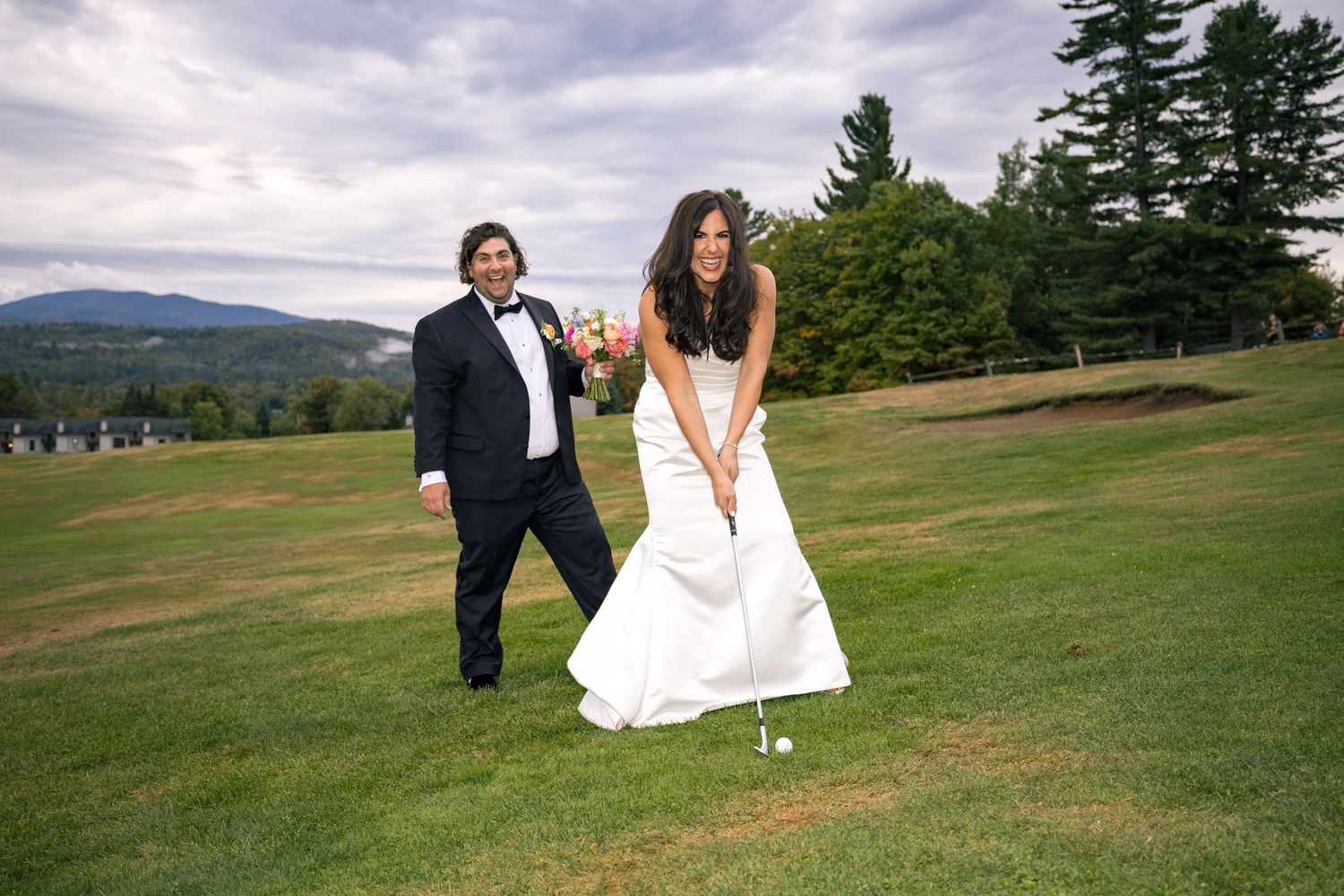 Bride in white gown playing golf while groom watches on a scenic green course with mountain views.