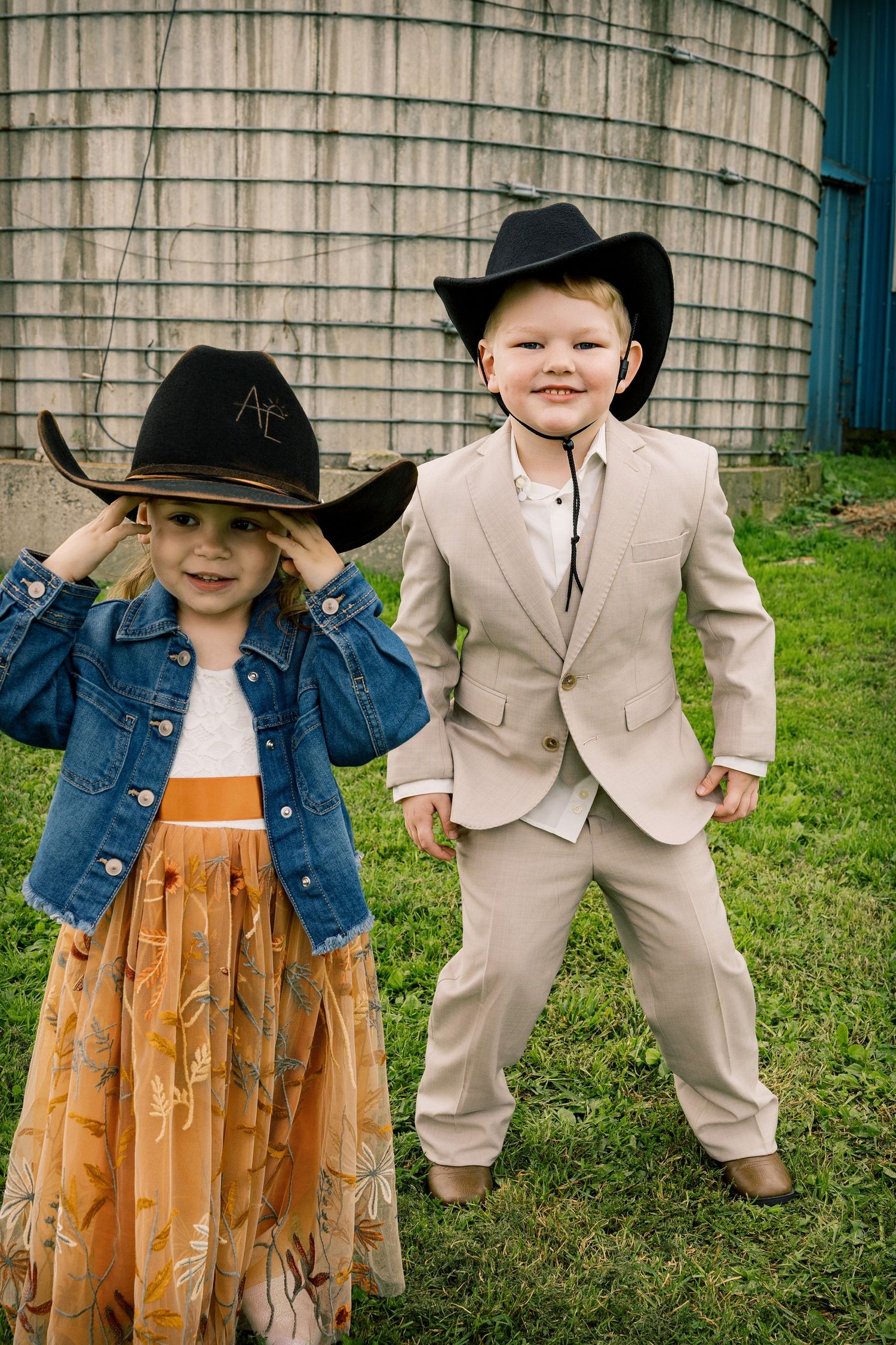 Two smiling children in black cowboy hats pose on the grass by a silo, a girl in a denim jacket and a boy in a tan suit.