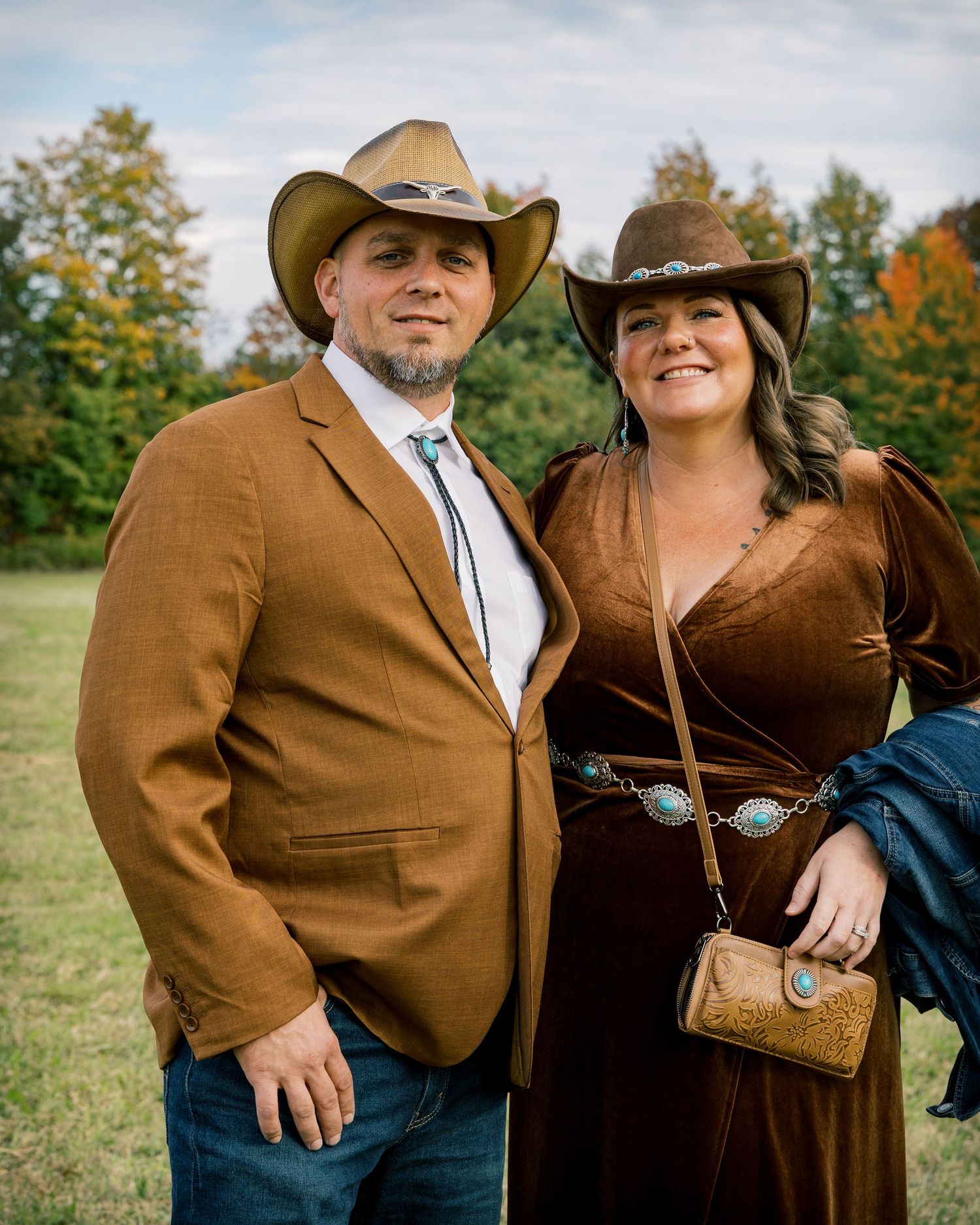 Smiling couple in matching brown cowboy hats, he in a rust blazer and she in a velvet dress, posing outdoors among autumn trees.