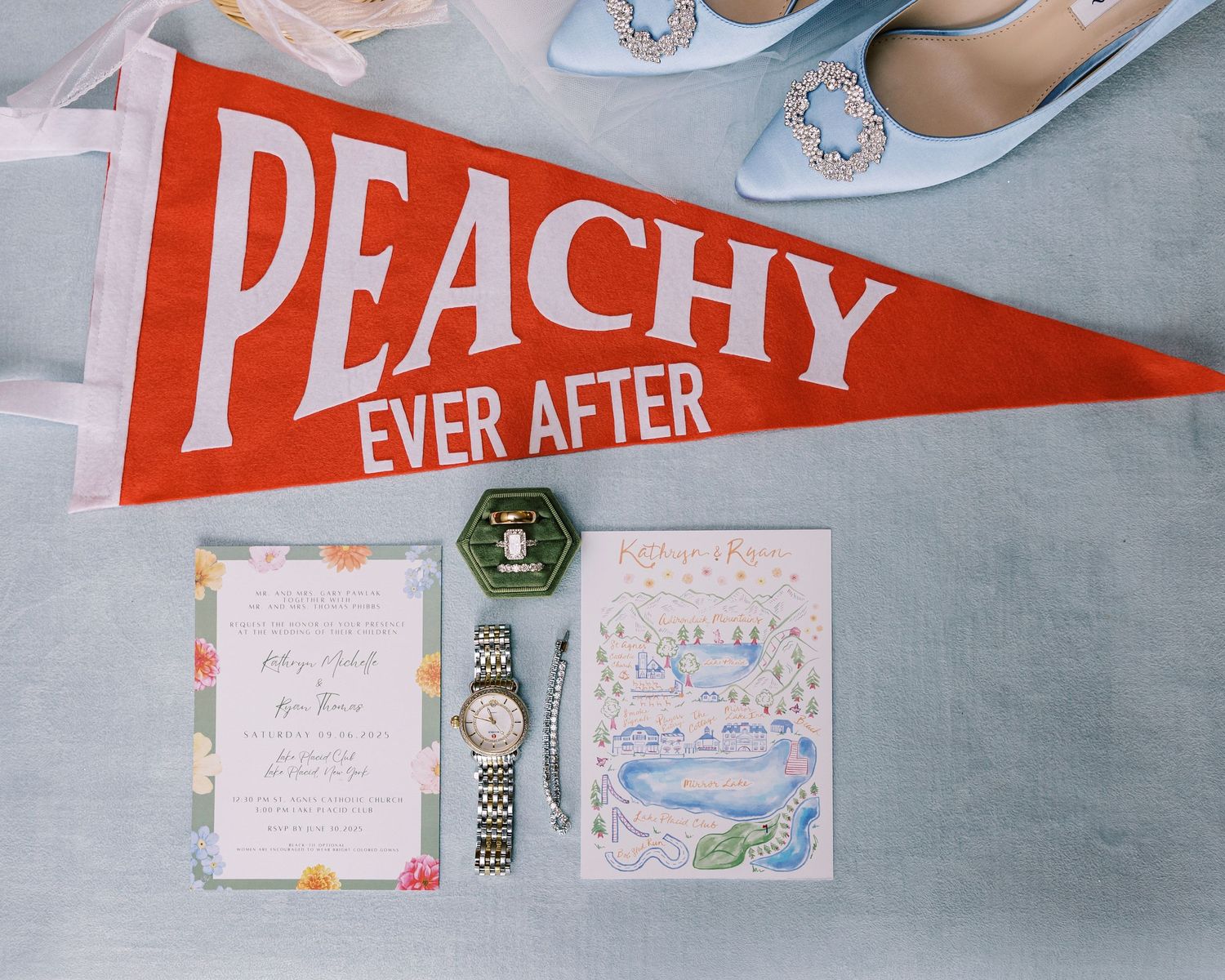 Wedding flat lay with Peachy Ever After pennant, floral invitations, ring box, watch, and blue heels on gray background.