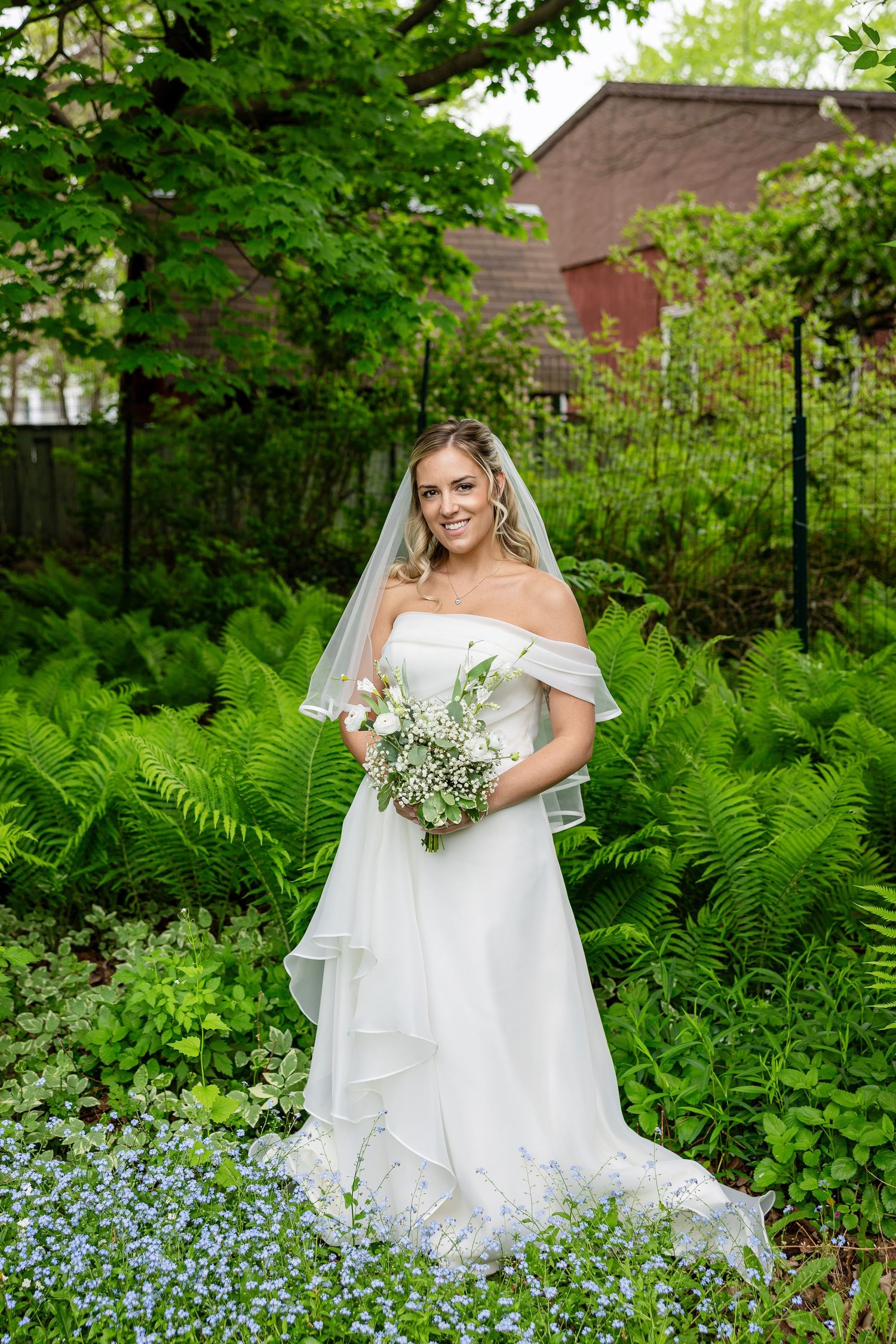 Bride in white off-shoulder gown and veil holds bouquet surrounded by lush green ferns and blue flowers outdoors.