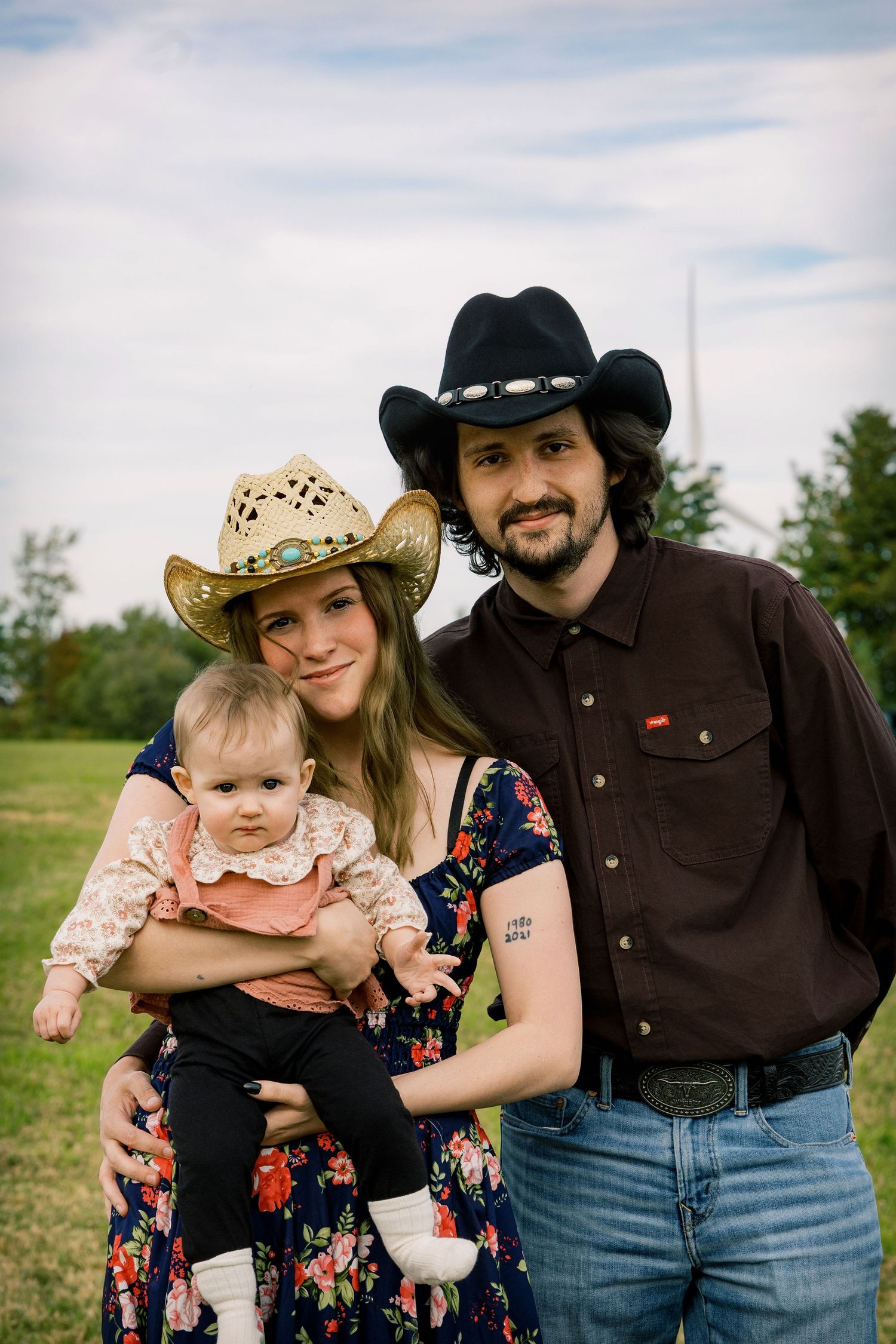 A smiling family in cowboy hats poses outdoors on a grassy field, with a woman in a floral dress holding a baby beside a bearded man.