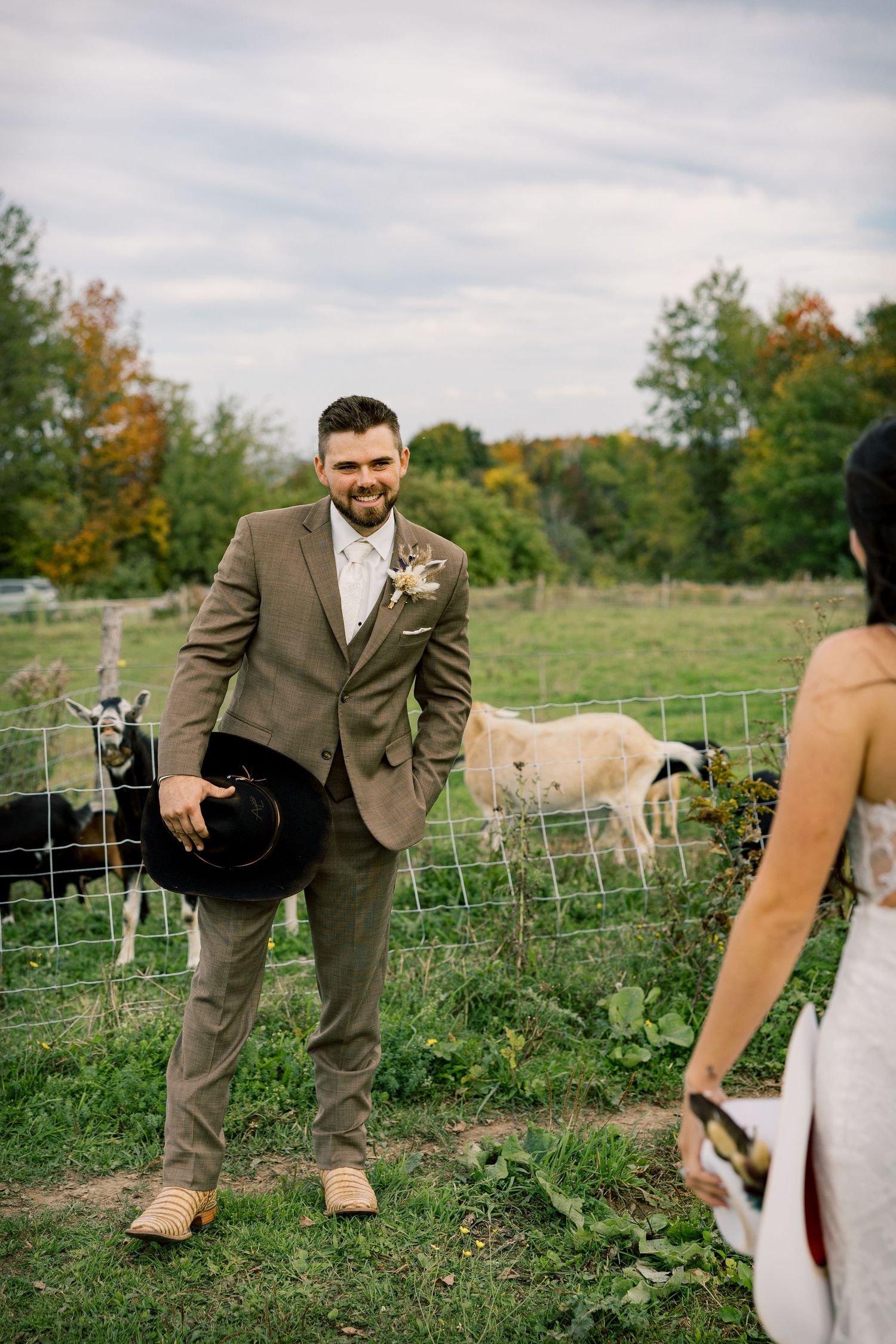 Smiling groom in a tan suit holds a black hat beside a goat pen on a farm while a bride in a white gown faces him.