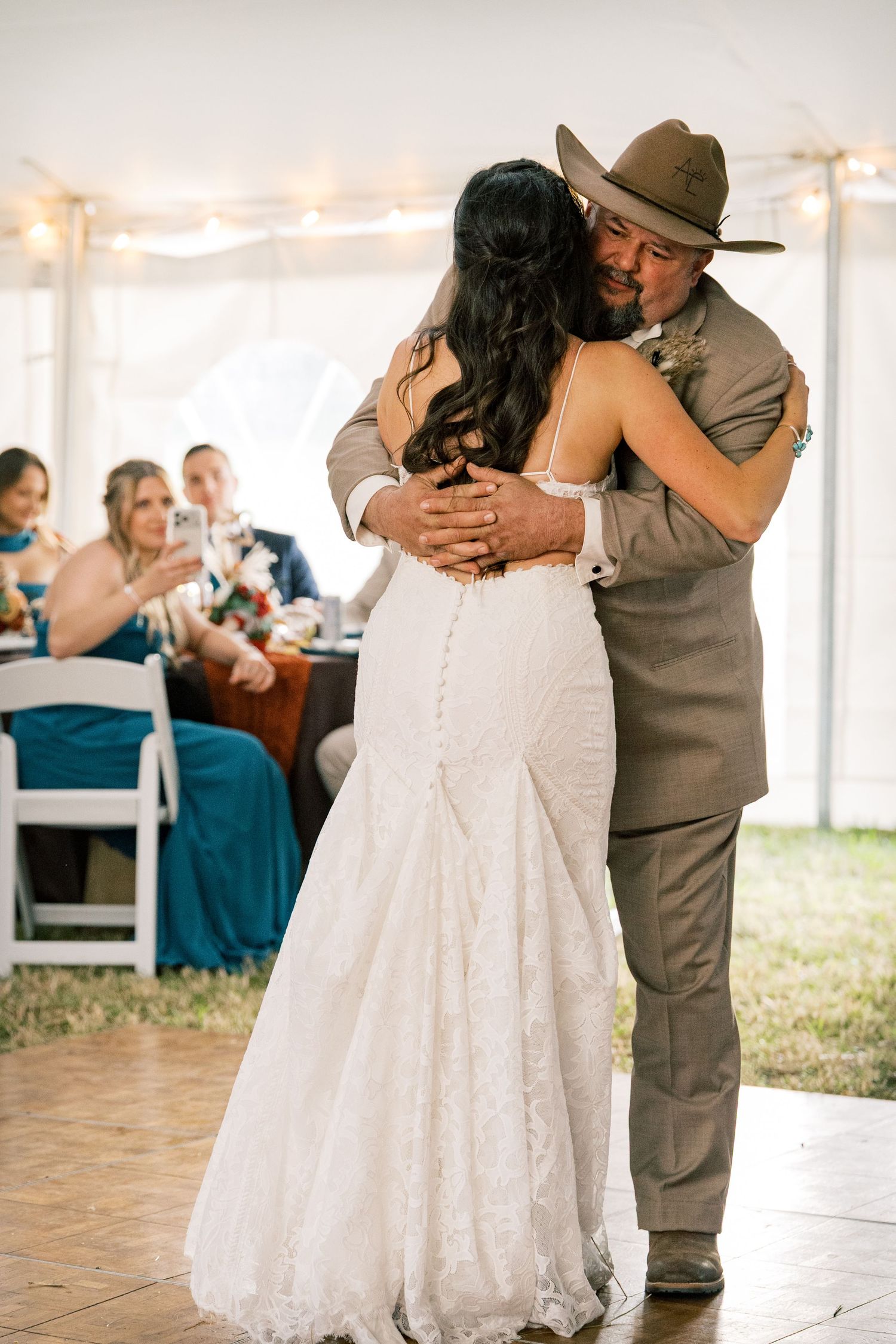 A bride in a lace gown dances with a man in a tan suit and cowboy hat while wedding guests watch from a tented reception.