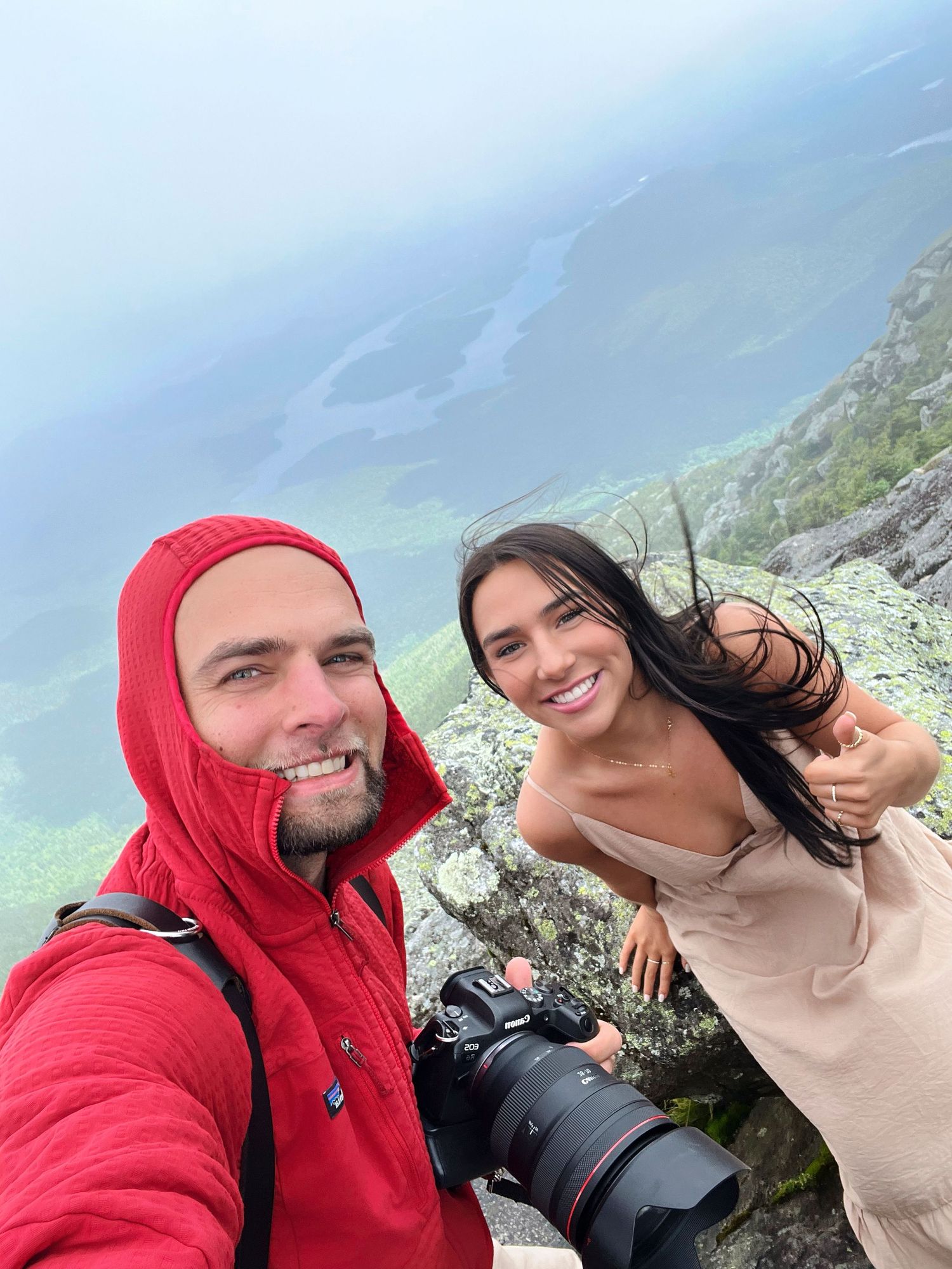 A couple takes selfies together while hiking in foggy mountain terrain wearing red and beige jackets.