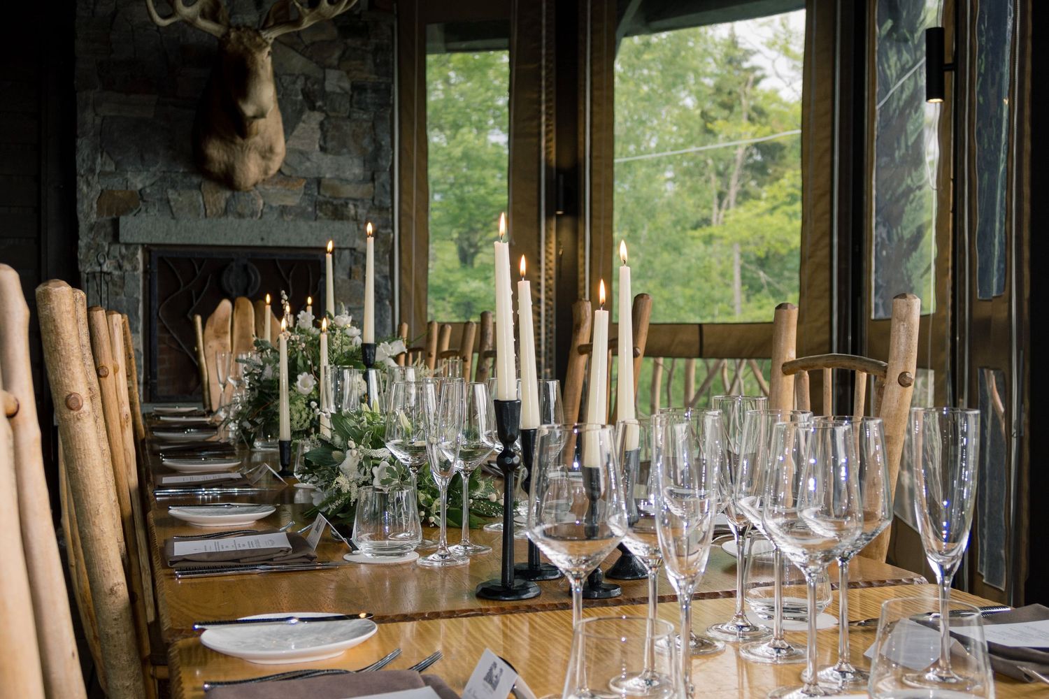 Rustic dining table set with tall candles, wine glasses, and greenery centerpieces in an elegant lodge with fireplace.