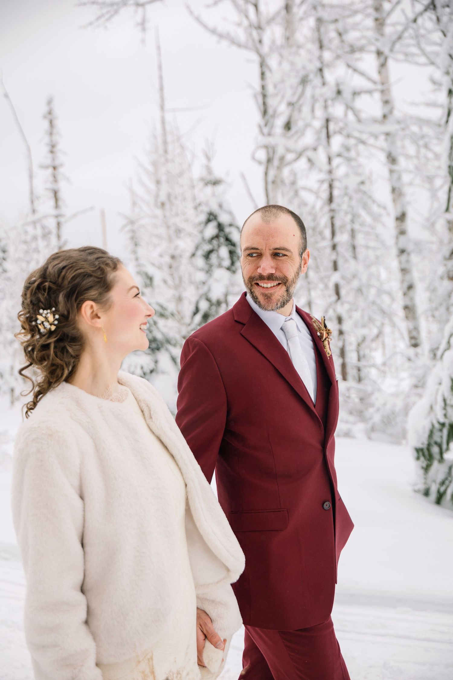 A couple shares joyful laughter together in a snowy winter forest setting while wearing burgundy and white formal attire.