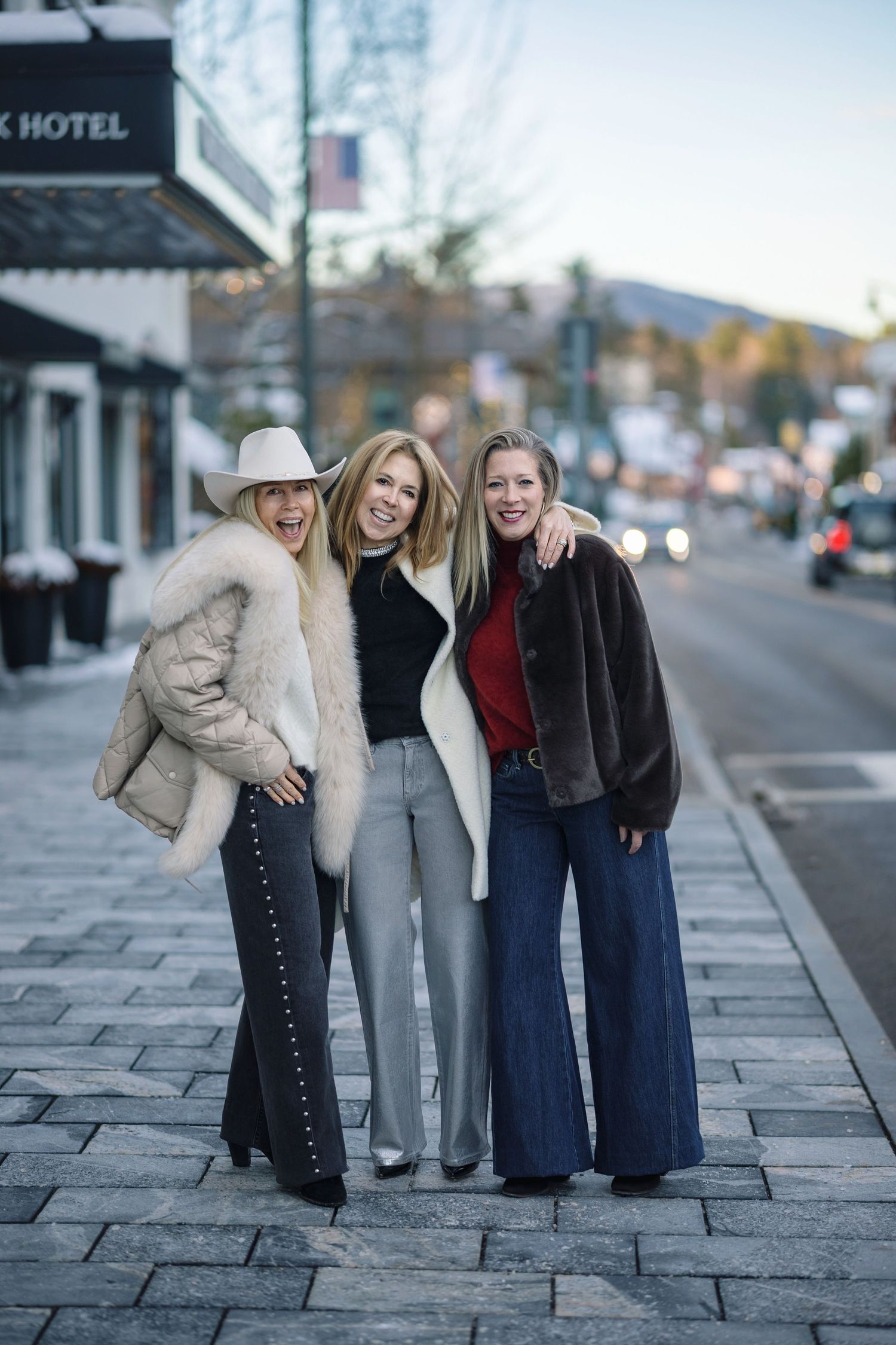 Three friends in winter coats and jeans pose together on a city sidewalk with storefronts in the background.