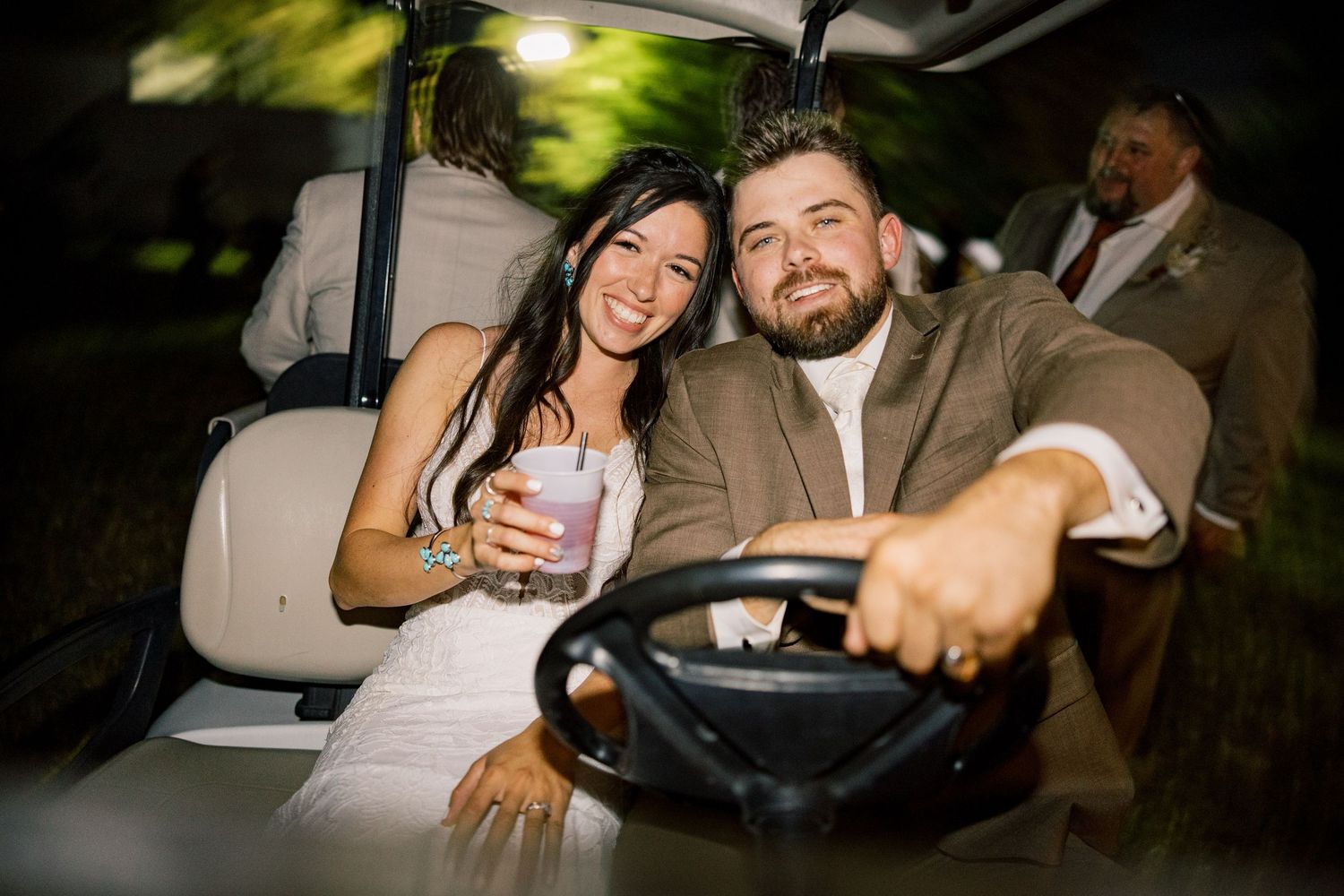 Smiling couple in formal wedding attire ride a golf cart at night, the woman holding a drink while the bearded man drives.