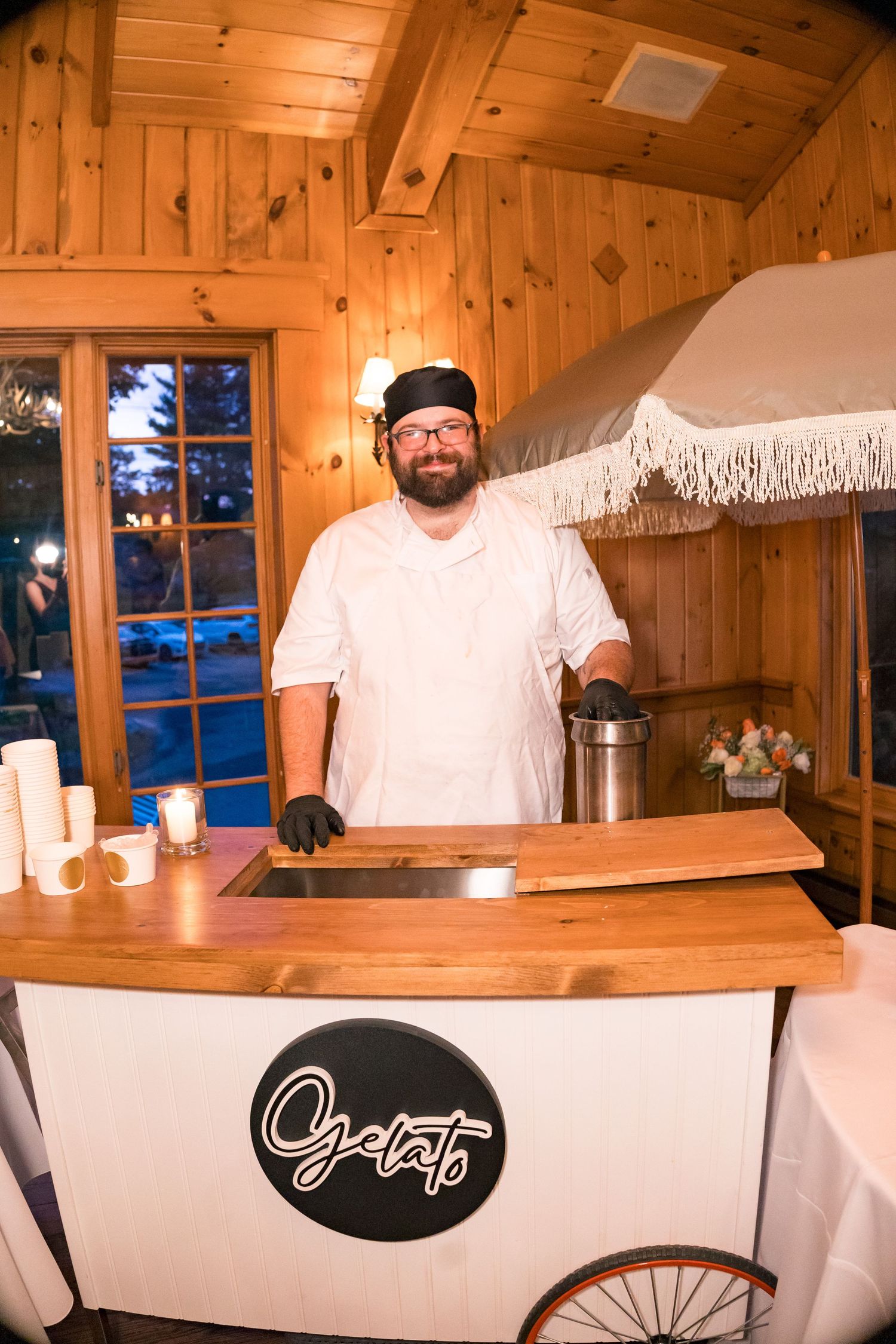 Smiling gelato vendor in white chef uniform stands behind a wooden gelato cart inside a warm wooden venue.