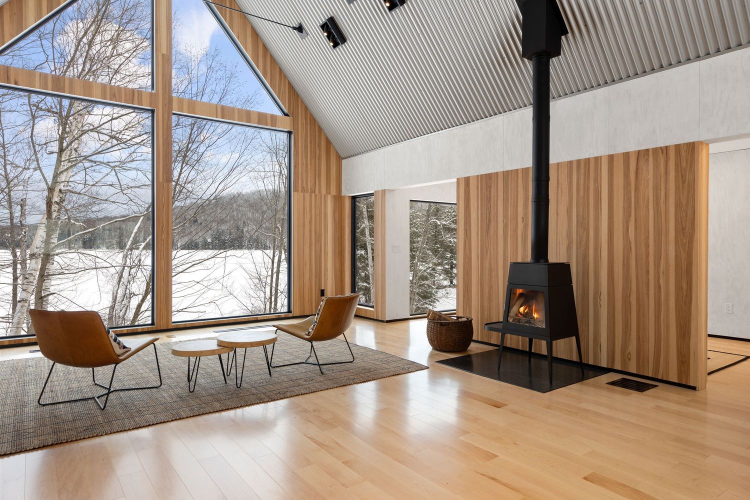 Modern cabin interior with large windows, wood panels, and a black wood stove overlooking a snowy landscape.