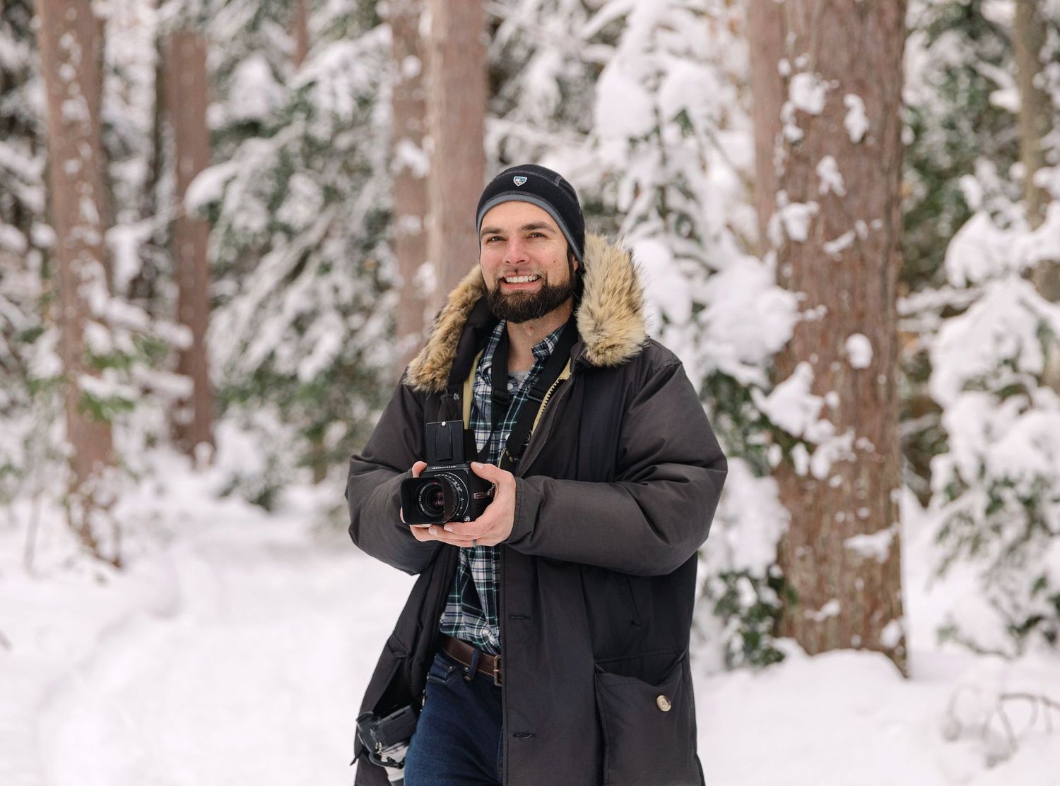 A photographer wearing a winter coat and beanie holds a camera while standing in a snowy forest.