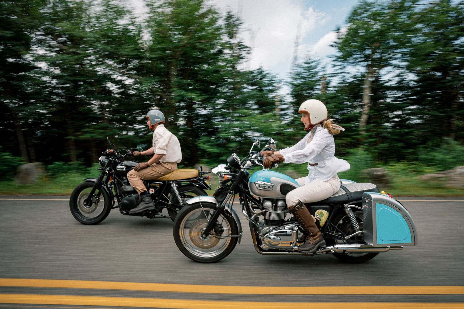 Motorcyclists riding vintage cafe racer bikes in formation on a tree-lined road during daytime.