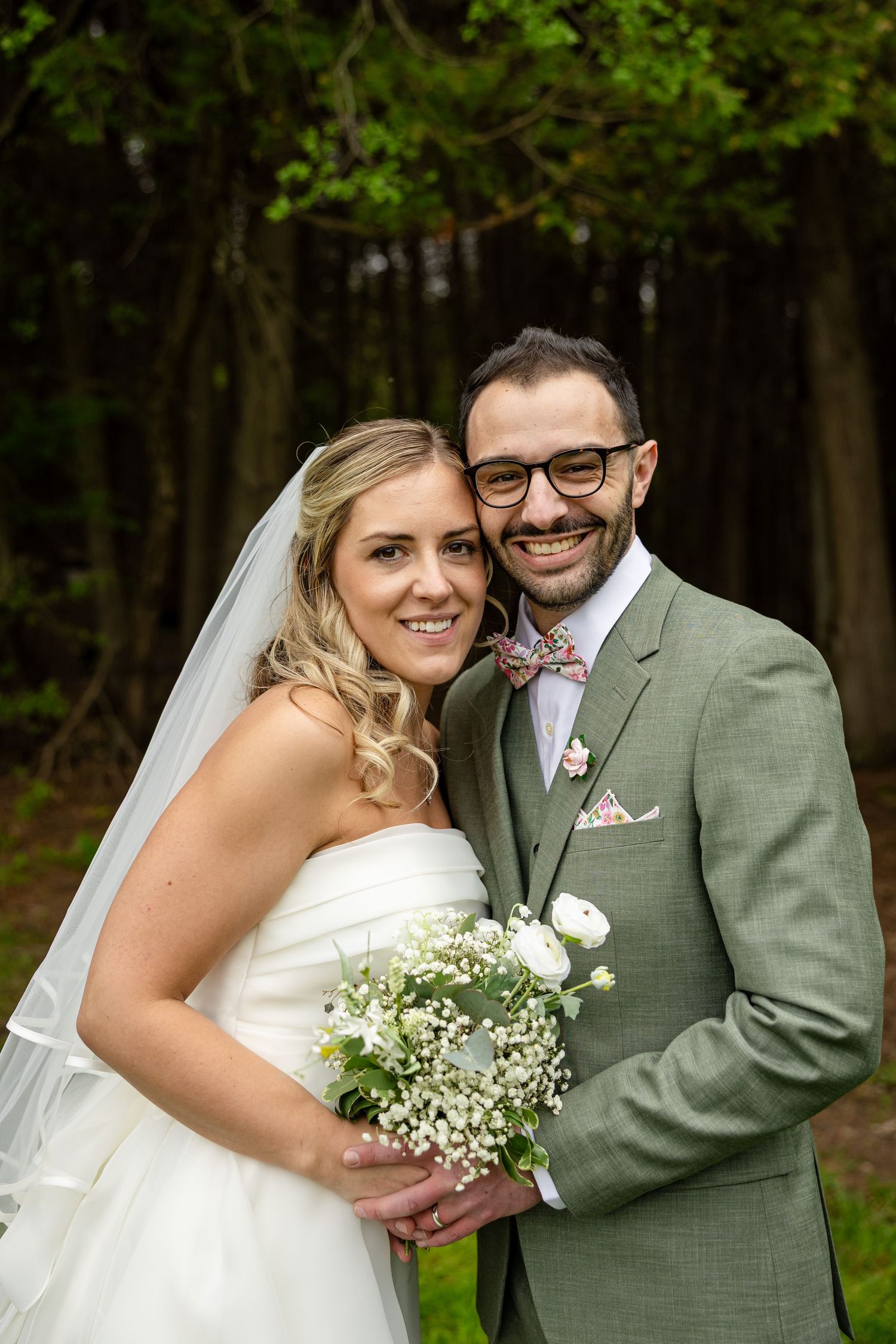 Smiling bride in white dress holding bouquet with groom in sage green suit and bow tie posing outdoors at wedding.