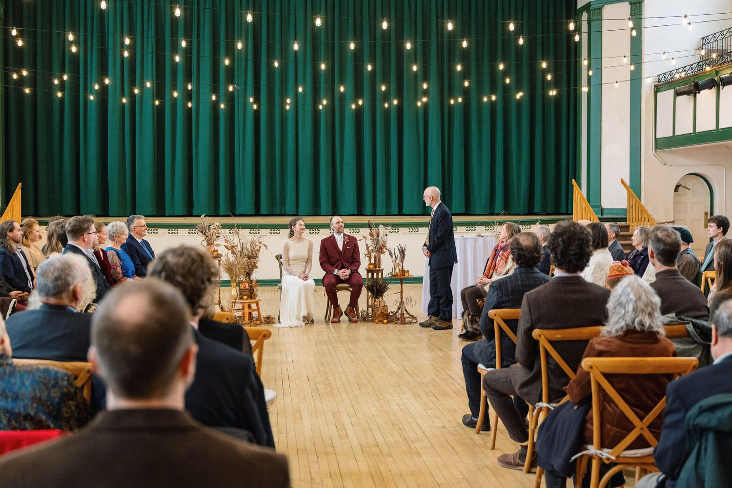 Guests seated in wooden chairs at an indoor event venue with string lights and green curtains along the walls.