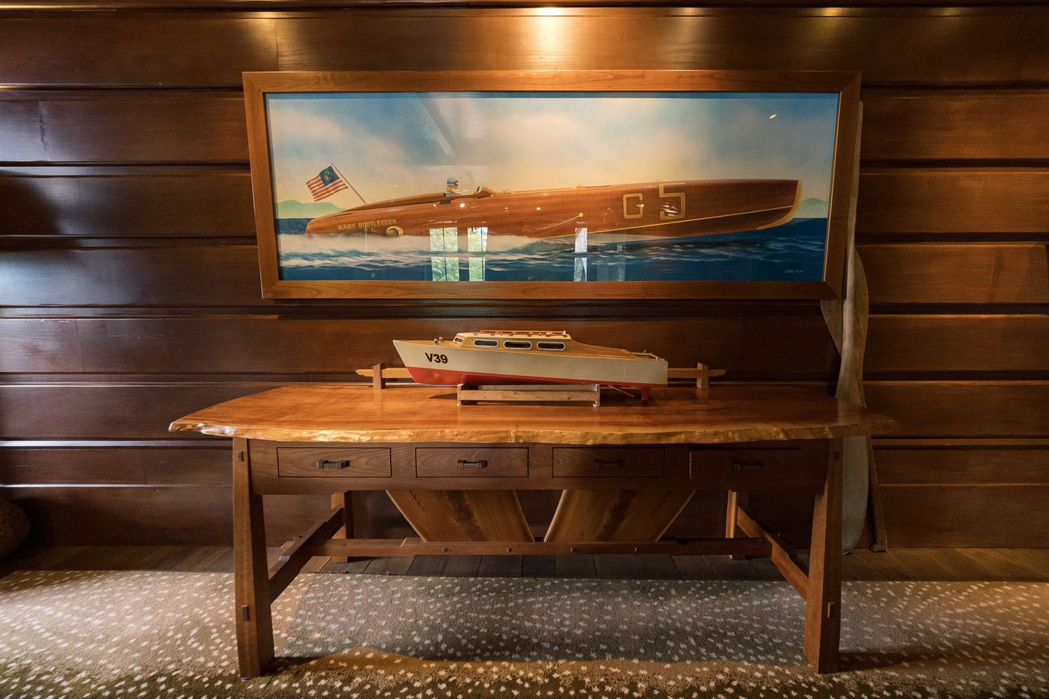 Wooden desk with model boat display and framed speedboat painting on wood-paneled wall in nautical decor setting.