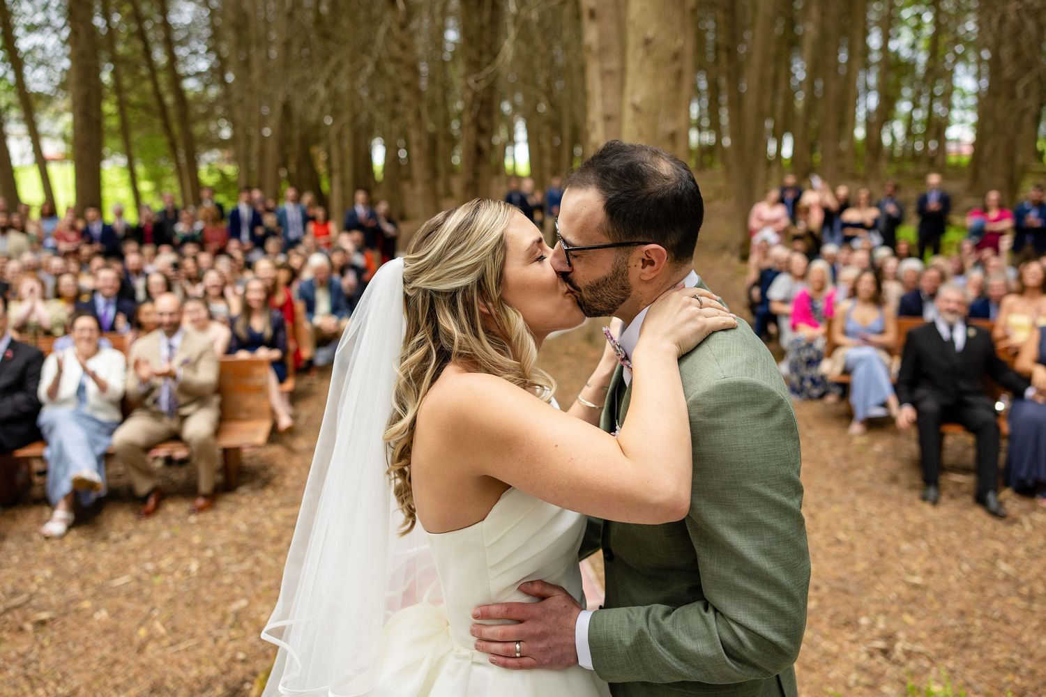 Bride and groom share first kiss at outdoor woodland wedding ceremony with guests seated among trees.