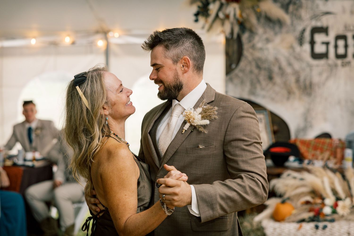A smiling woman dances closely with a bearded man in a tan suit and floral boutonniere at a rustic tented wedding reception.