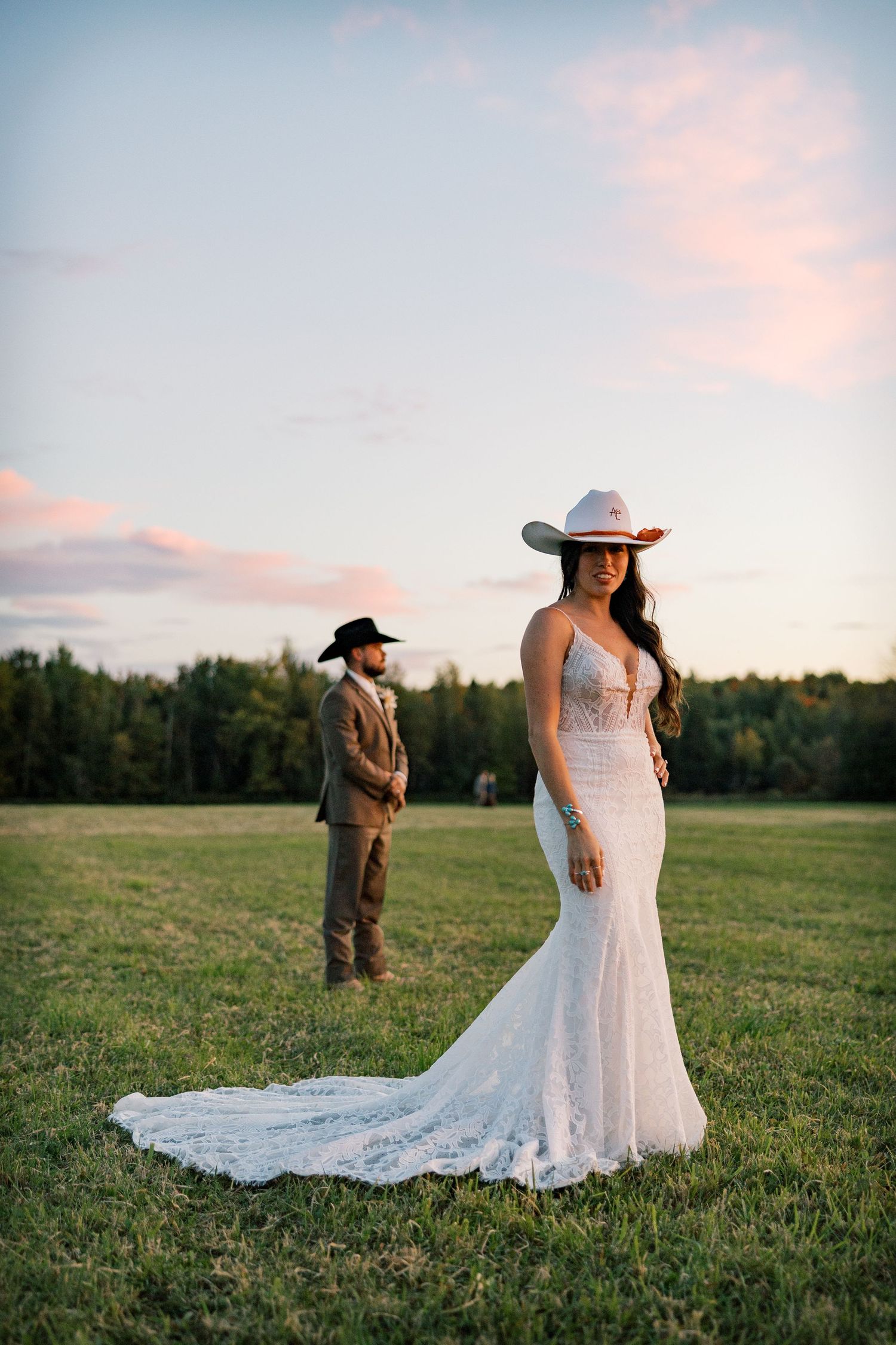 Bride in a lace mermaid gown and cowboy hat stands in a field at sunset as the groom in a western hat watches behind.
