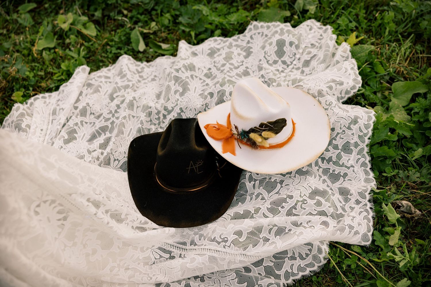 Black and white cowboy hats with feather and floral accents rest on a white lace veil spread across green grass.