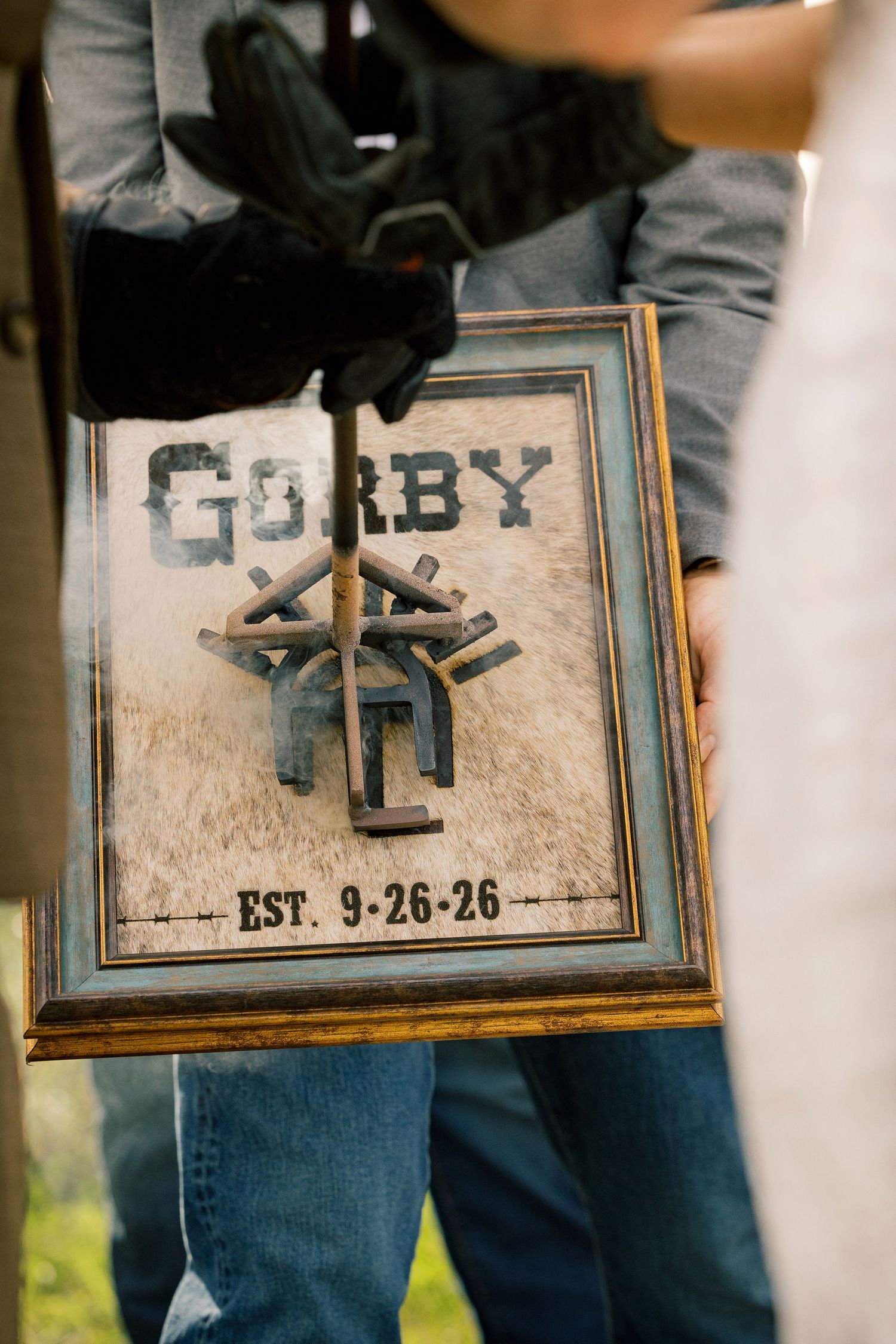 Gloved hand holds a branding iron over a framed Gorby sign on cowhide, marked Est. 9-26-26, in a rustic western wedding ceremony.