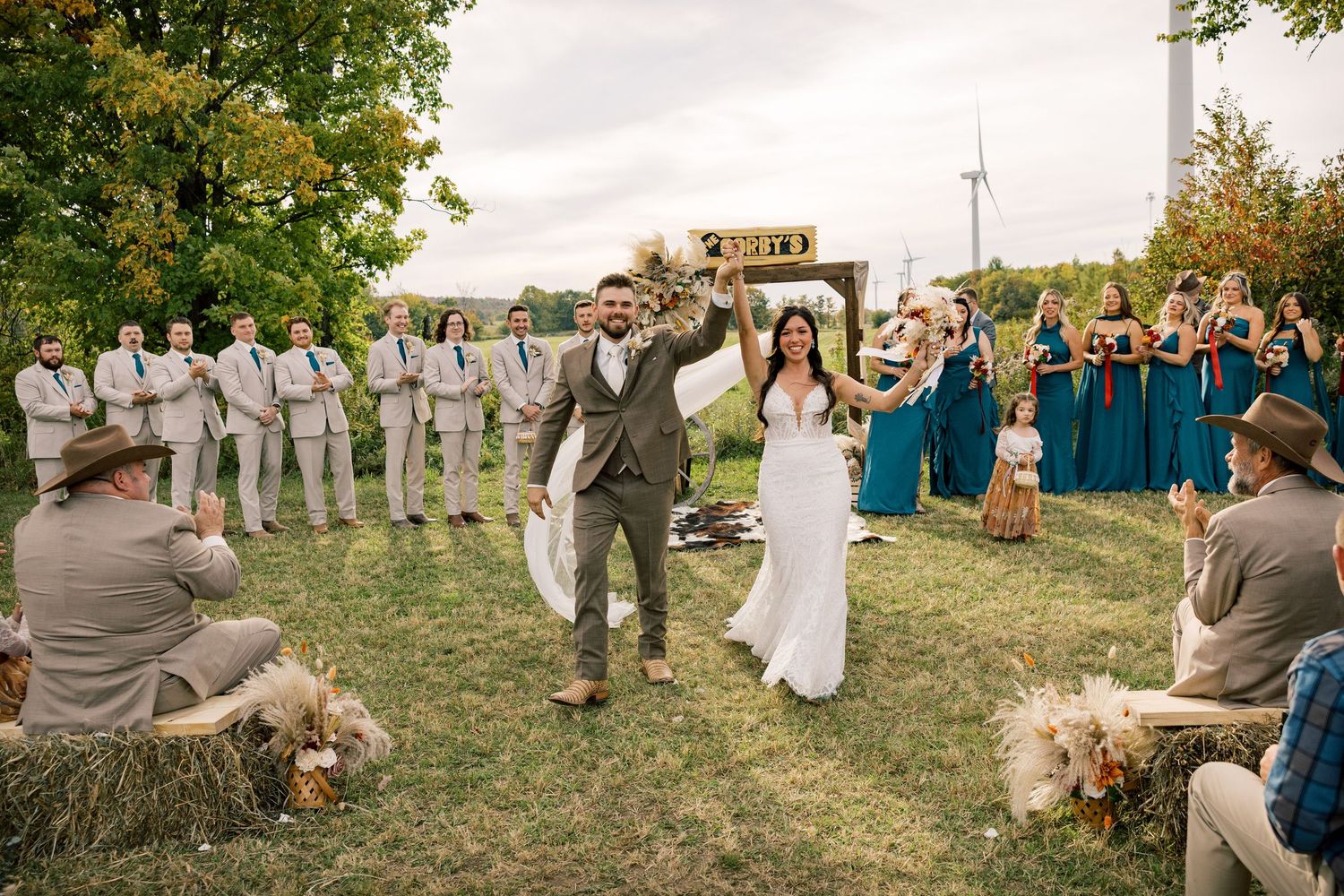 Bride and groom raise their arms in celebration at an outdoor farm wedding ceremony, with groomsmen, bridesmaids and guests on hay bales.