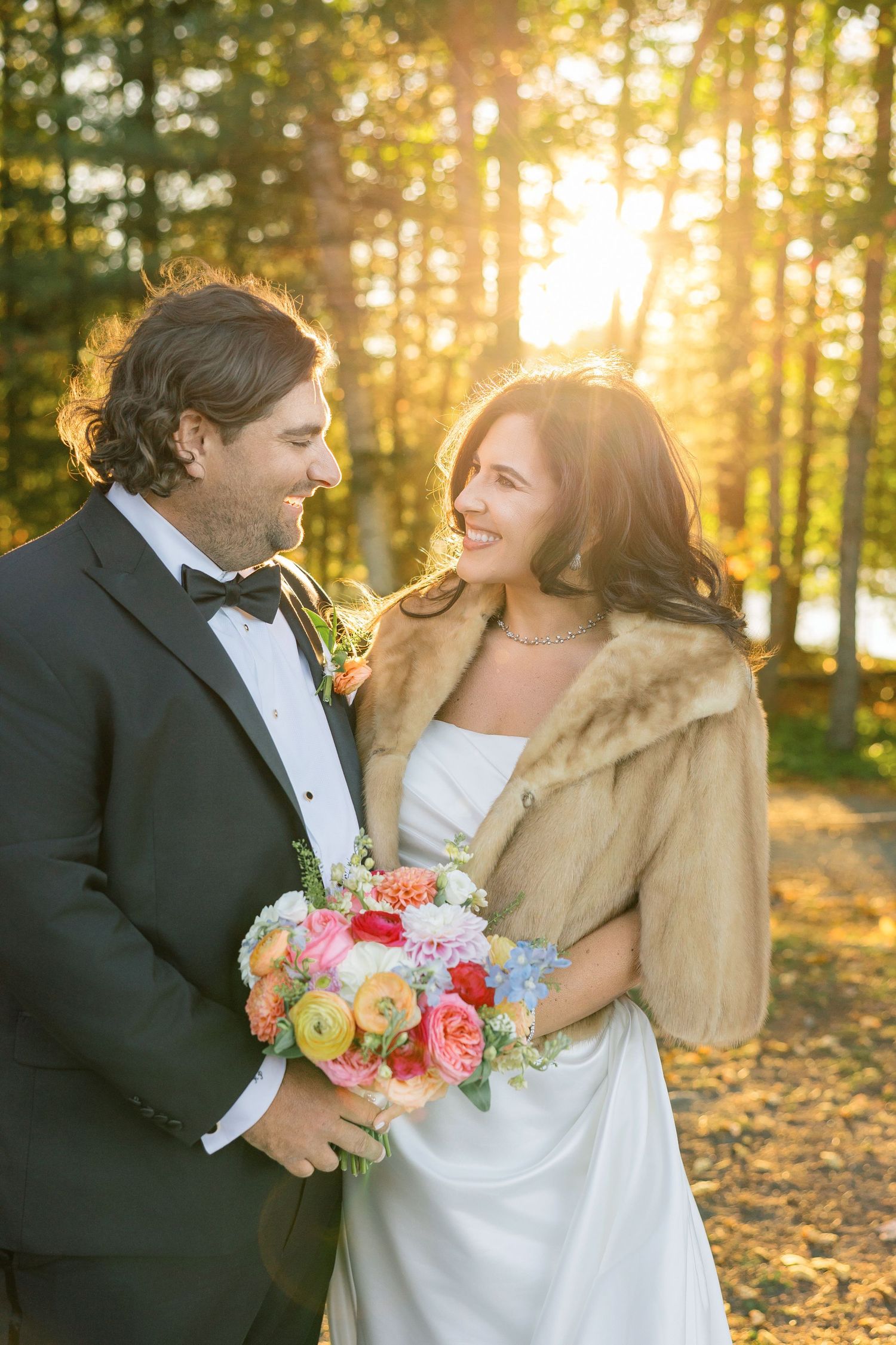 Bride and groom smiling at each other in golden sunlight, she wears fur stole and holds colorful bouquet.