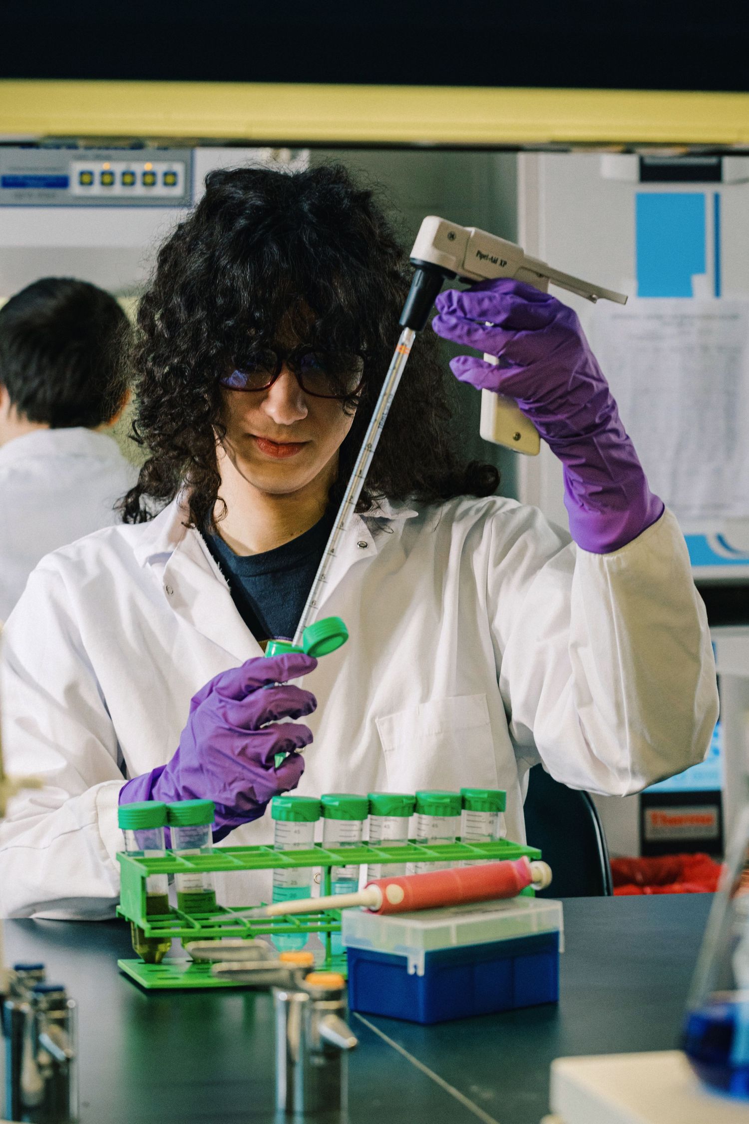 Laboratory technician in white coat and purple gloves examines green scientific equipment during research experiment.