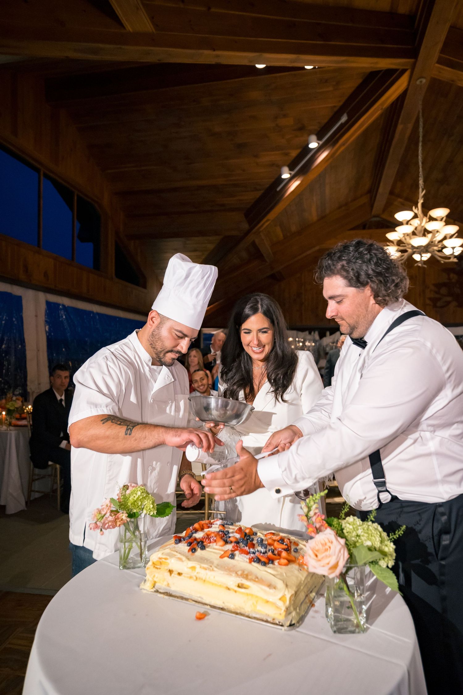 Chef and couple cutting a decorated wedding cake at an elegant indoor reception venue with wood beam ceiling.