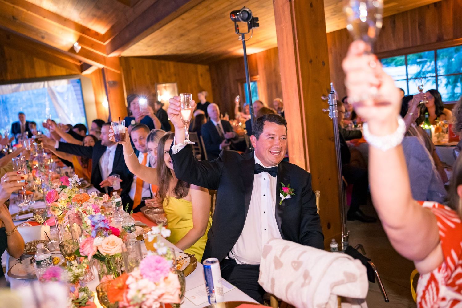 Wedding guests raise glasses in a toast at a elegant reception inside a rustic wood-paneled venue with floral centerpieces.