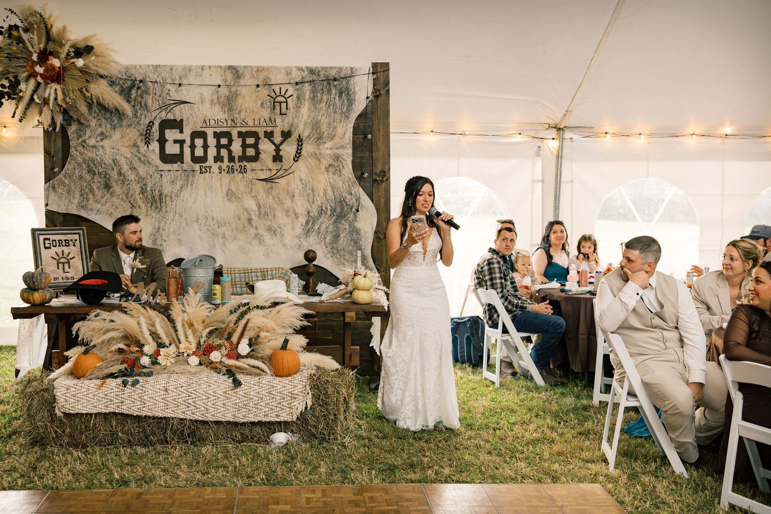 Bride in a white gown speaks into a microphone at a rustic tent wedding reception with guests seated and a Gorby sign behind her.