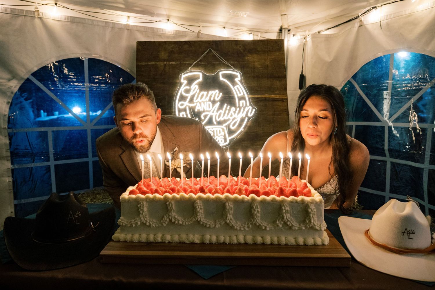 Couple smiling behind a large sheet cake covered in lit candles at a rustic party with a neon horseshoe sign and cowboy hats on the table.