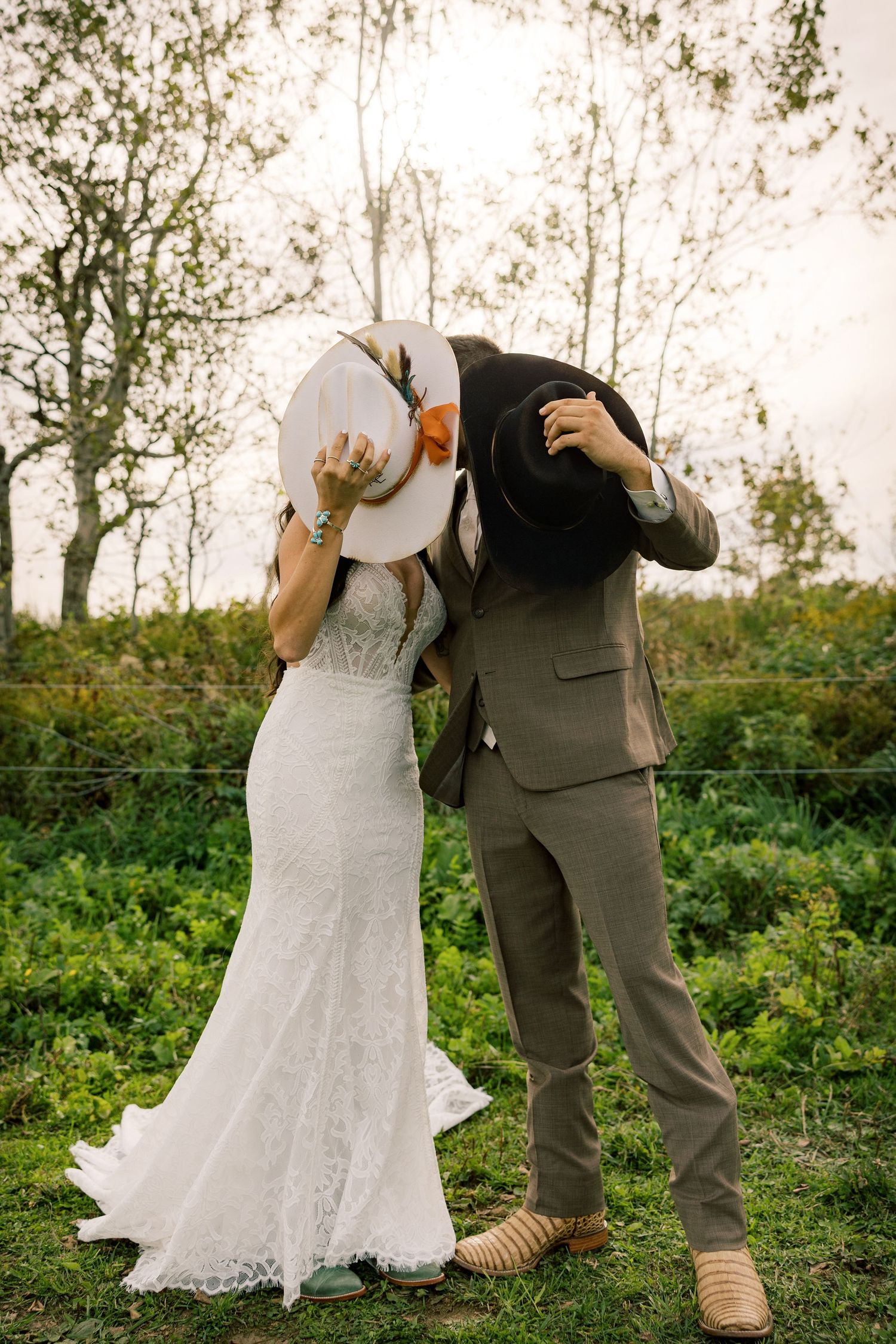 Bride in a lace wedding gown and groom in a tan suit hide their kiss behind cowboy hats in a green field.