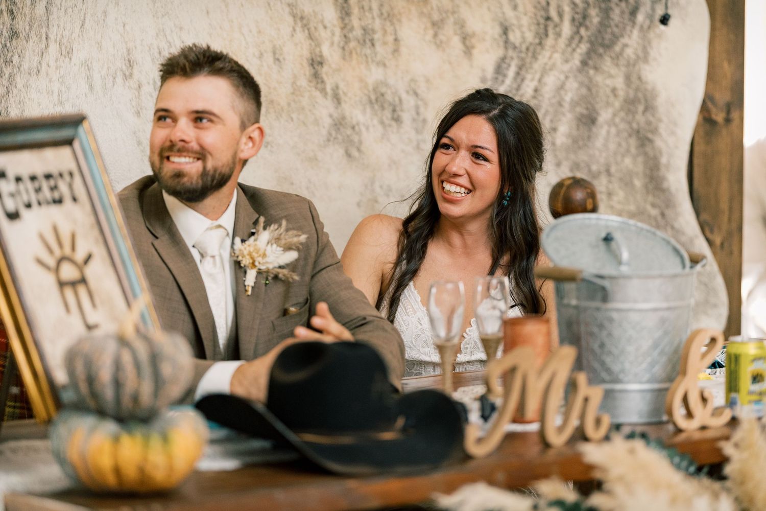 Smiling bride and groom seated at a rustic sweetheart table with a cowboy hat, pumpkins, wooden letters, and a metal ice bucket.