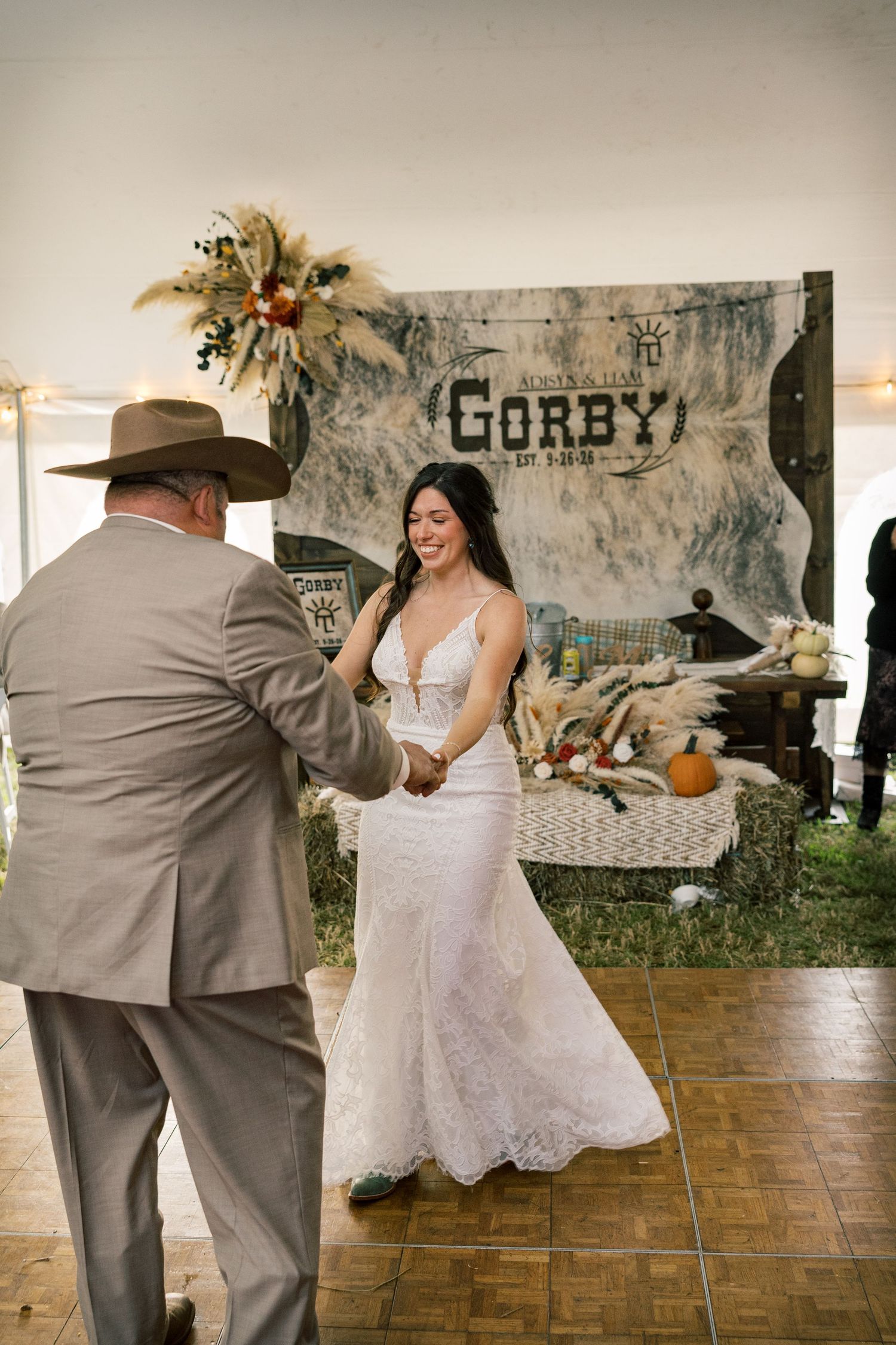 Bride in a fitted lace wedding dress dances with a man in a cowboy hat under a tent with western Gorby rustic decor.