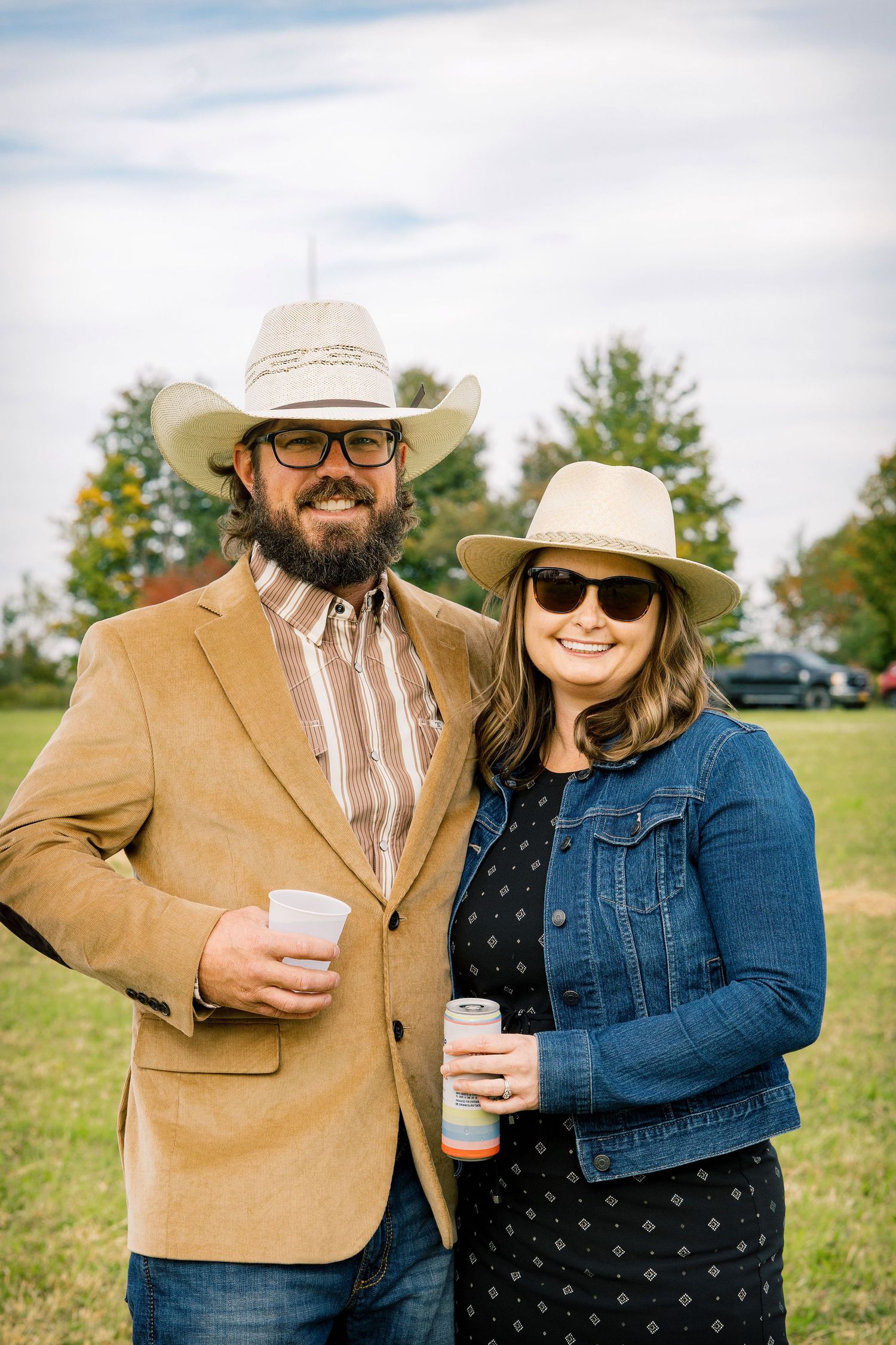 Smiling couple in cowboy hats and sunglasses hold drinks while posing together at an outdoor event in a grassy field.