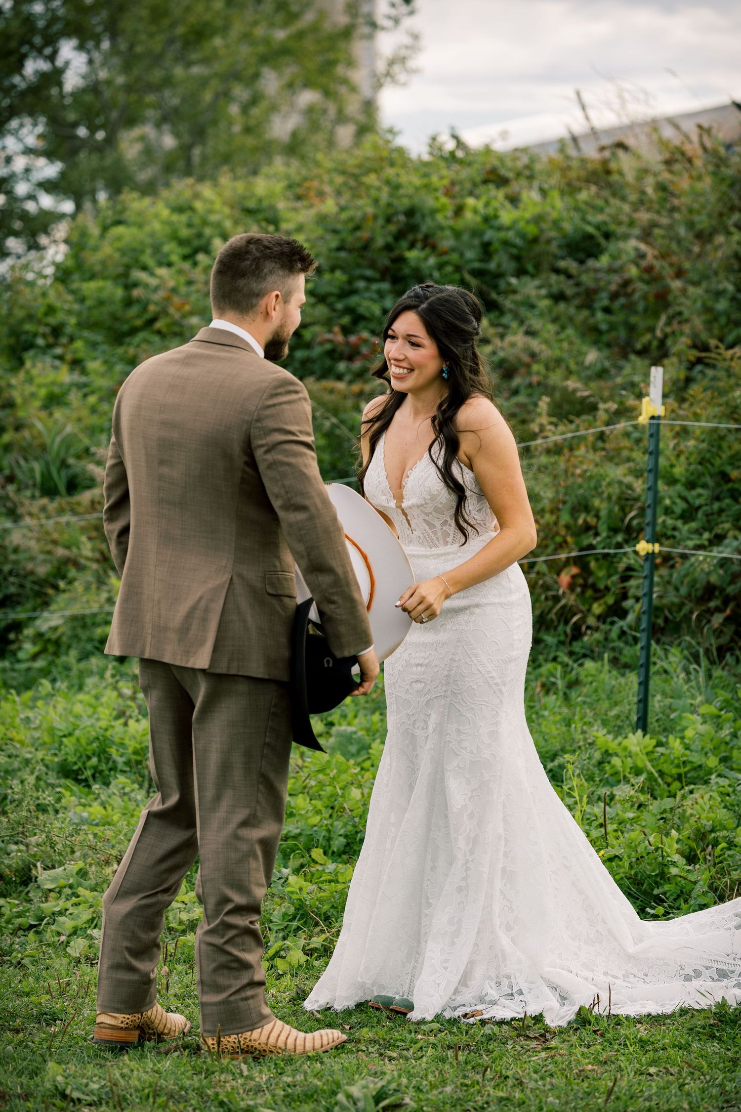 Smiling bride in a white lace wedding gown faces a groom in a brown tweed suit holding a hat during an outdoor first look.