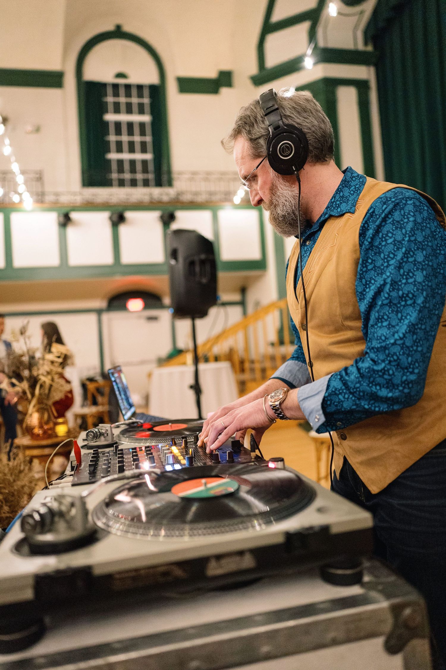 A DJ in a yellow vest and blue shirt operates turntables and mixing equipment during a performance.
