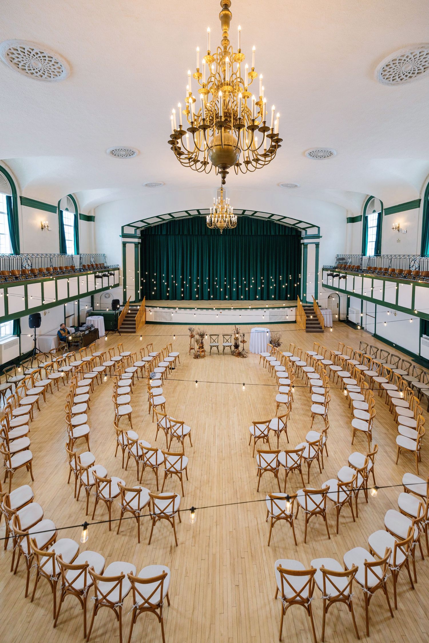 Elegant ballroom with wooden chairs arranged in circular pattern under ornate gold chandeliers and green-trimmed stage.