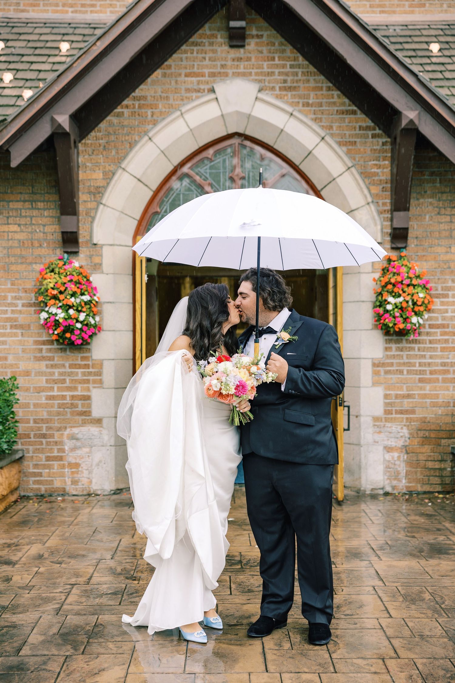 Bride and groom kissing under white umbrella outside brick church, bride holding colorful bouquet in wedding dress.