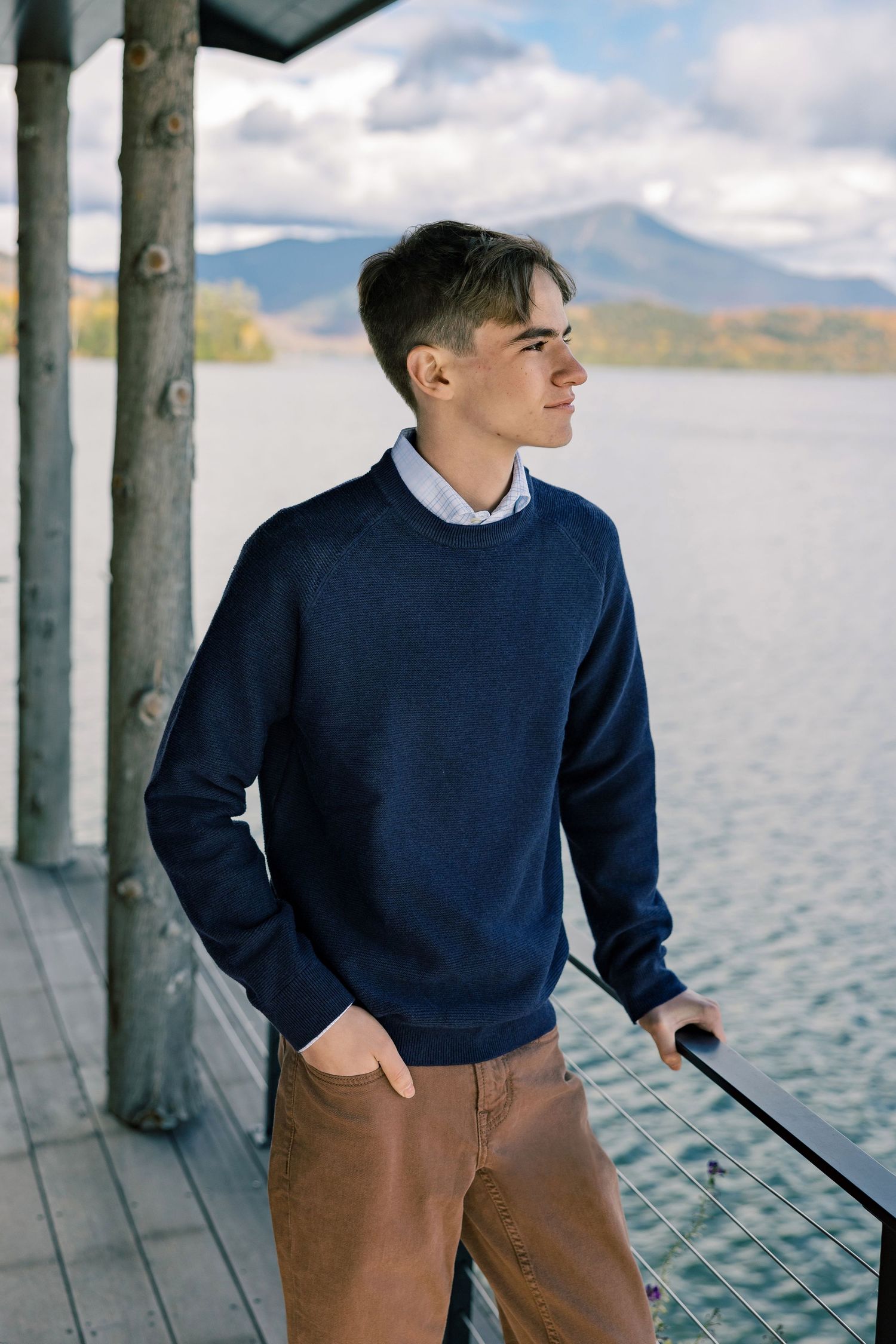 Man in navy sweater and brown pants stands on wooden pier overlooking serene lake with mountains in background.