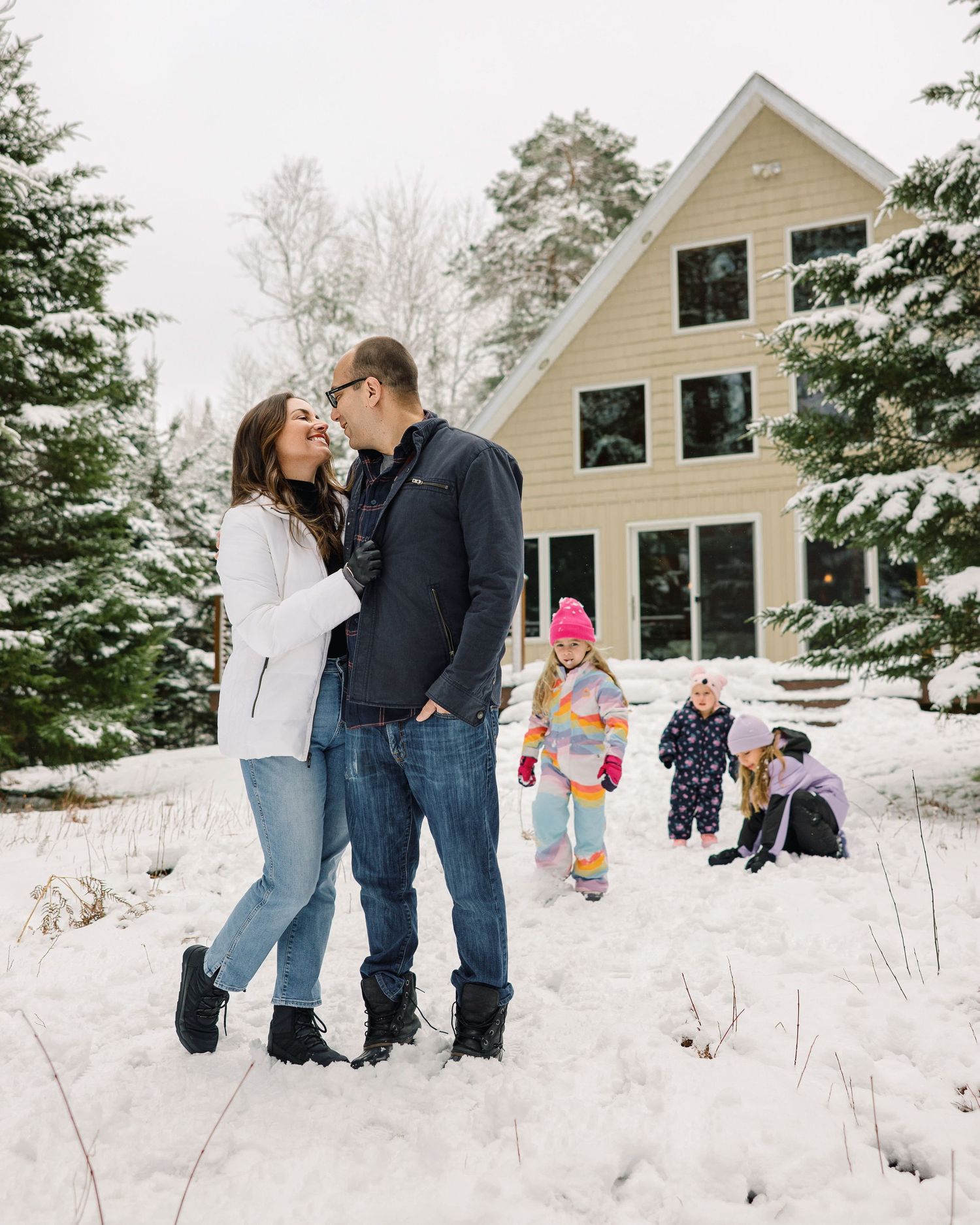A couple and children play together in a snowy winter landscape outside a beige house.