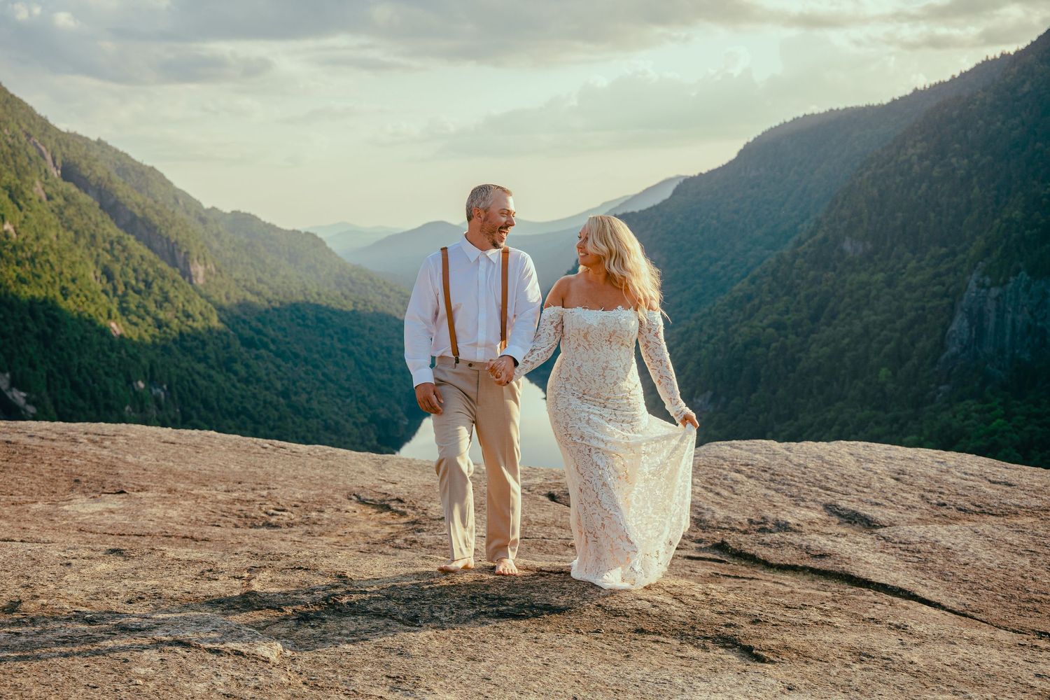 Couple in white attire walking along scenic mountain overlook at sunset.