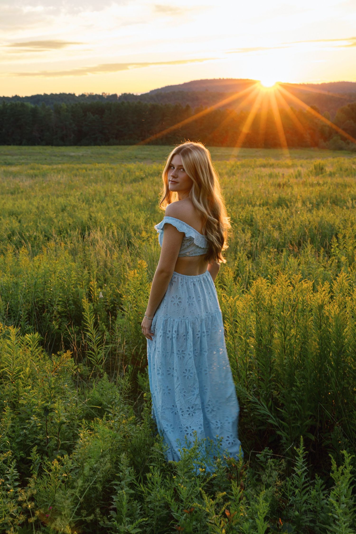 A person in a light blue dress stands in a golden meadow at sunset with sunbeams streaming through the mountains.