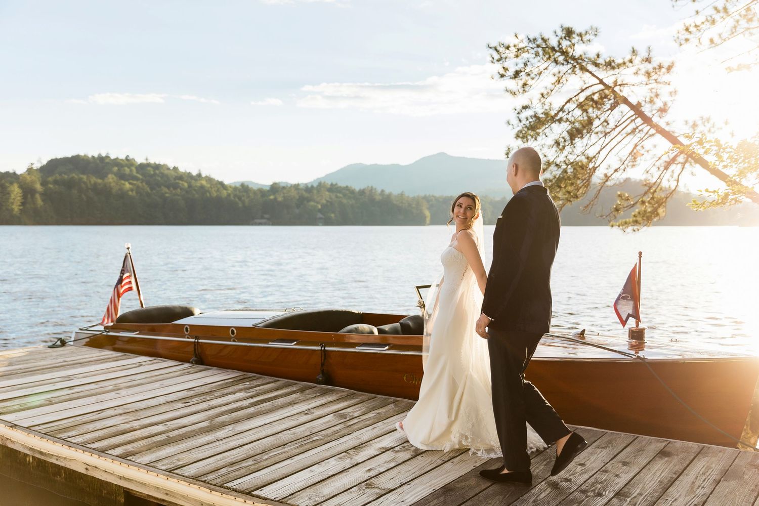 A couple in wedding attire stands on a wooden dock by a lake with a wooden boat at sunset.