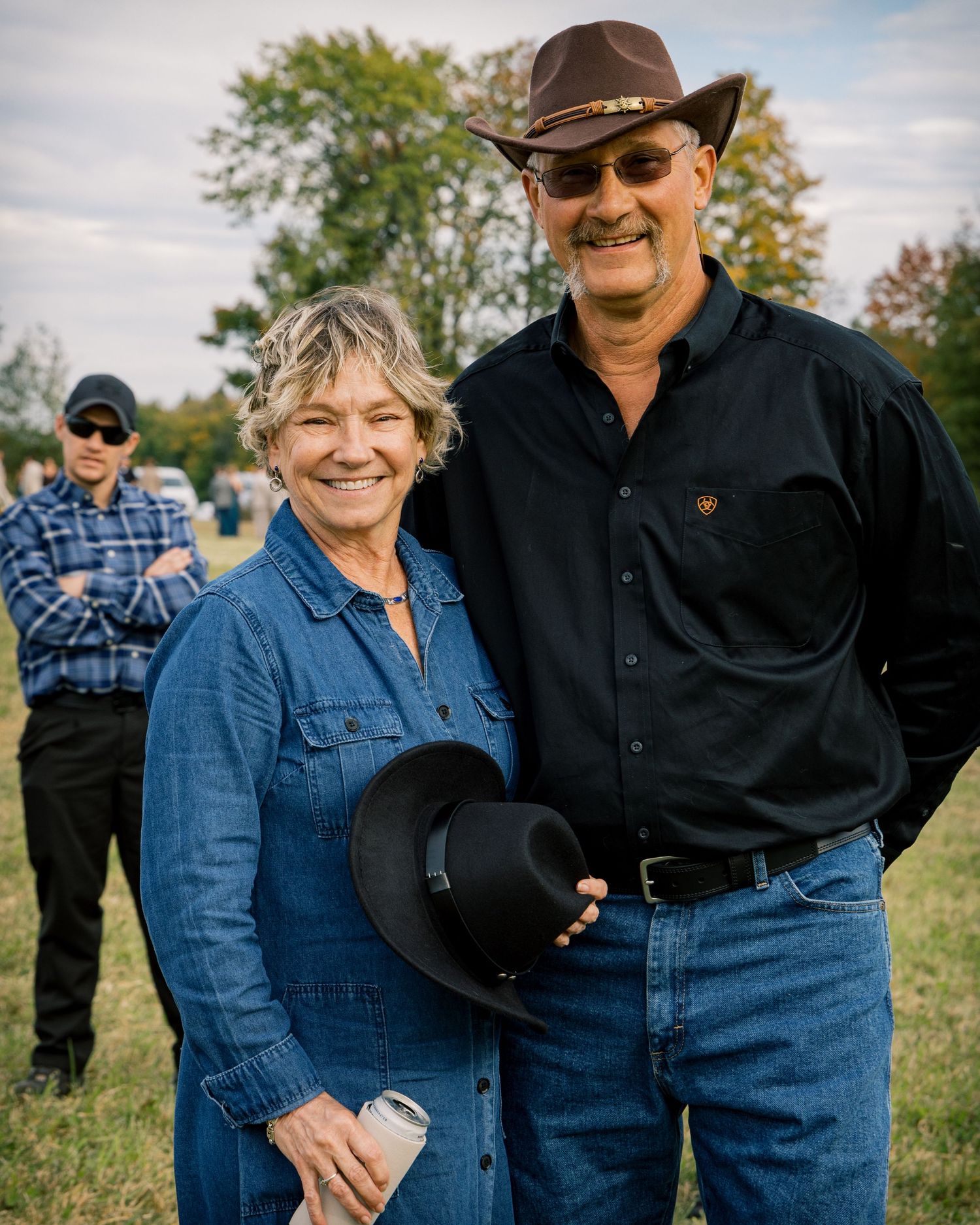 Smiling couple in denim and western wear pose together in a grassy field, the man in a brown hat and sunglasses, with a person behind them.