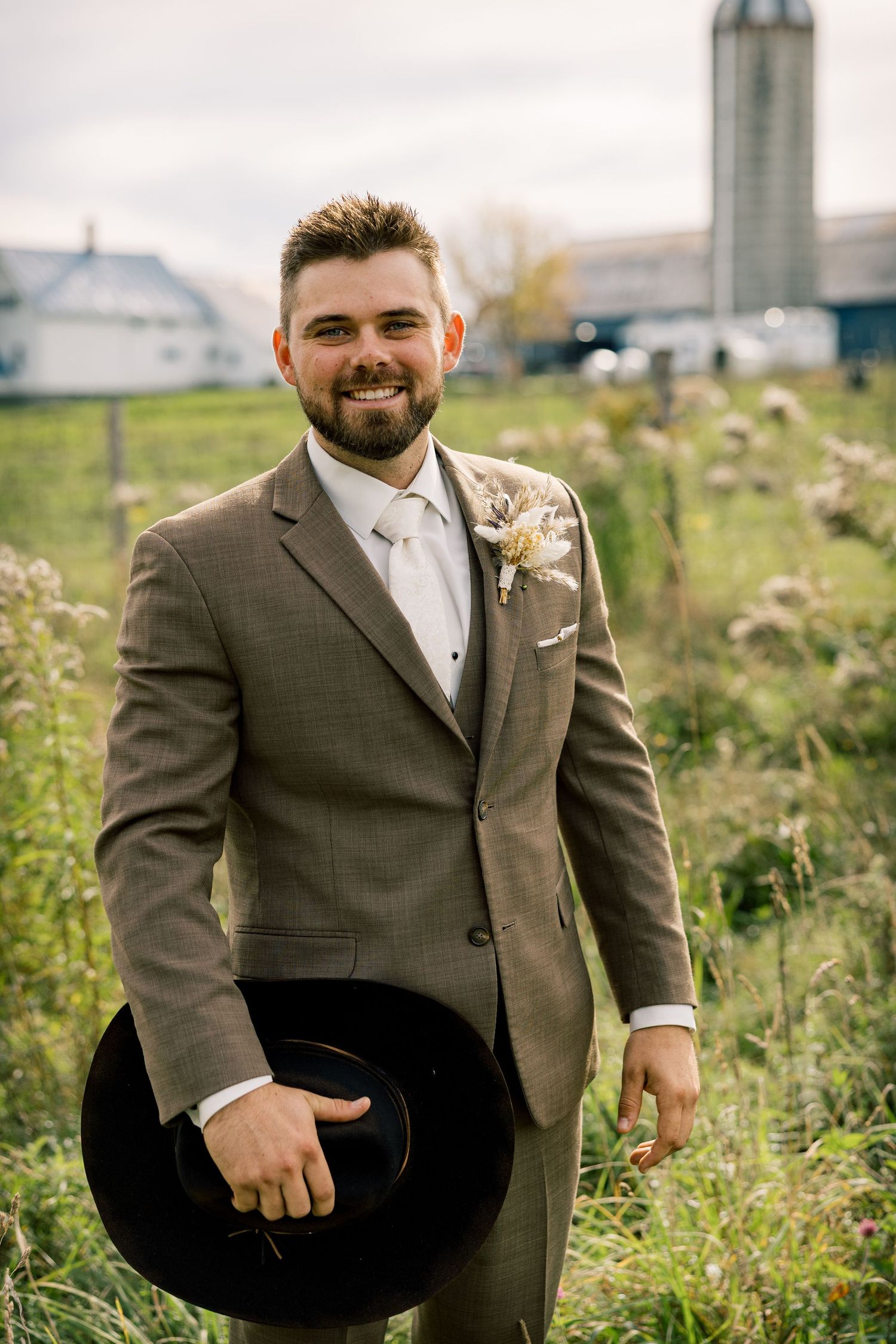 Smiling groom in a brown tweed suit with a dried floral boutonniere holds a black cowboy hat in a grassy field with a farm silo behind.